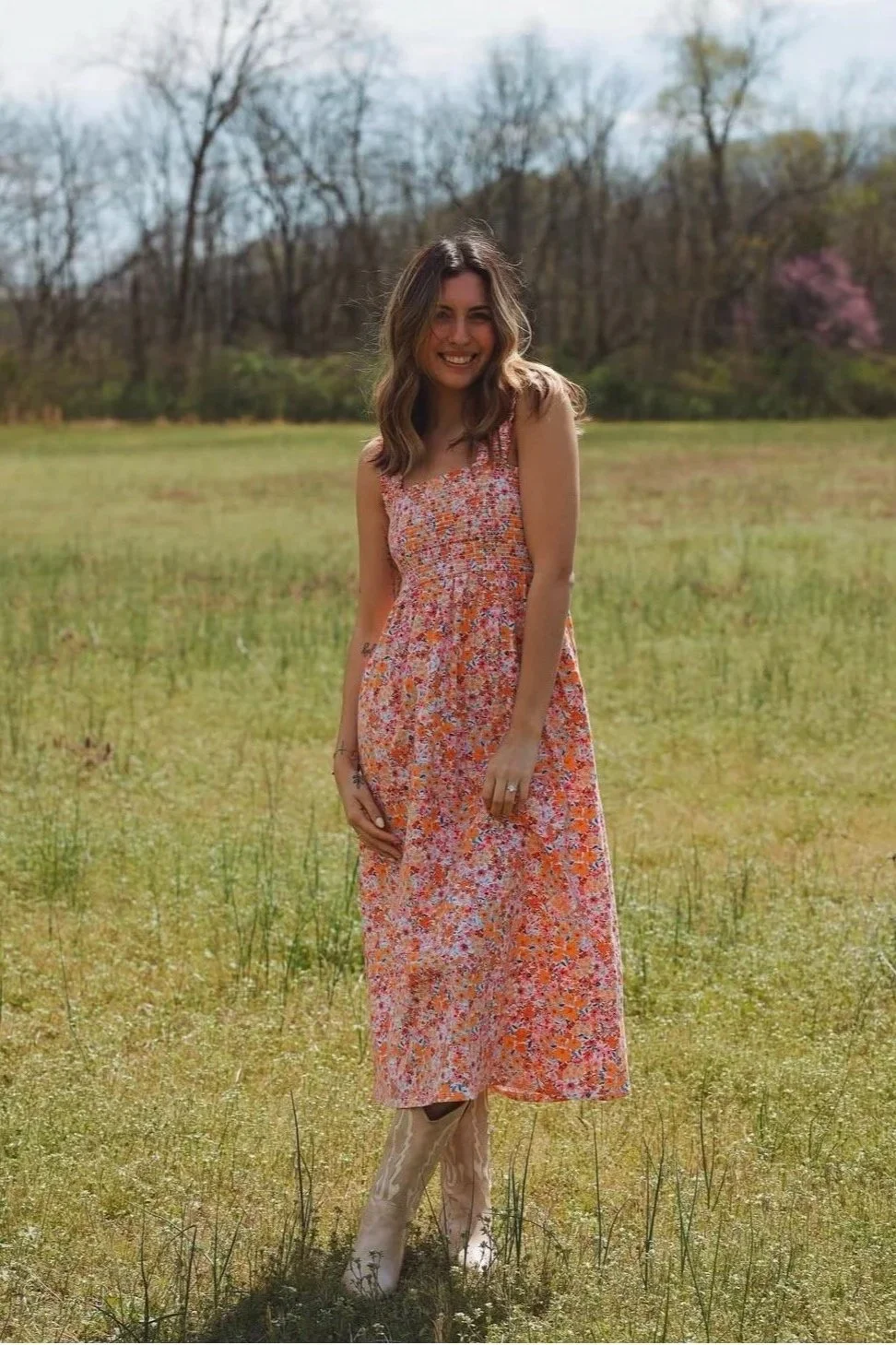 A young woman with brown hair wearing a sleeveless, floral, orange and pink dress, standing in a grassy field with trees and a hill in the background, smiling.