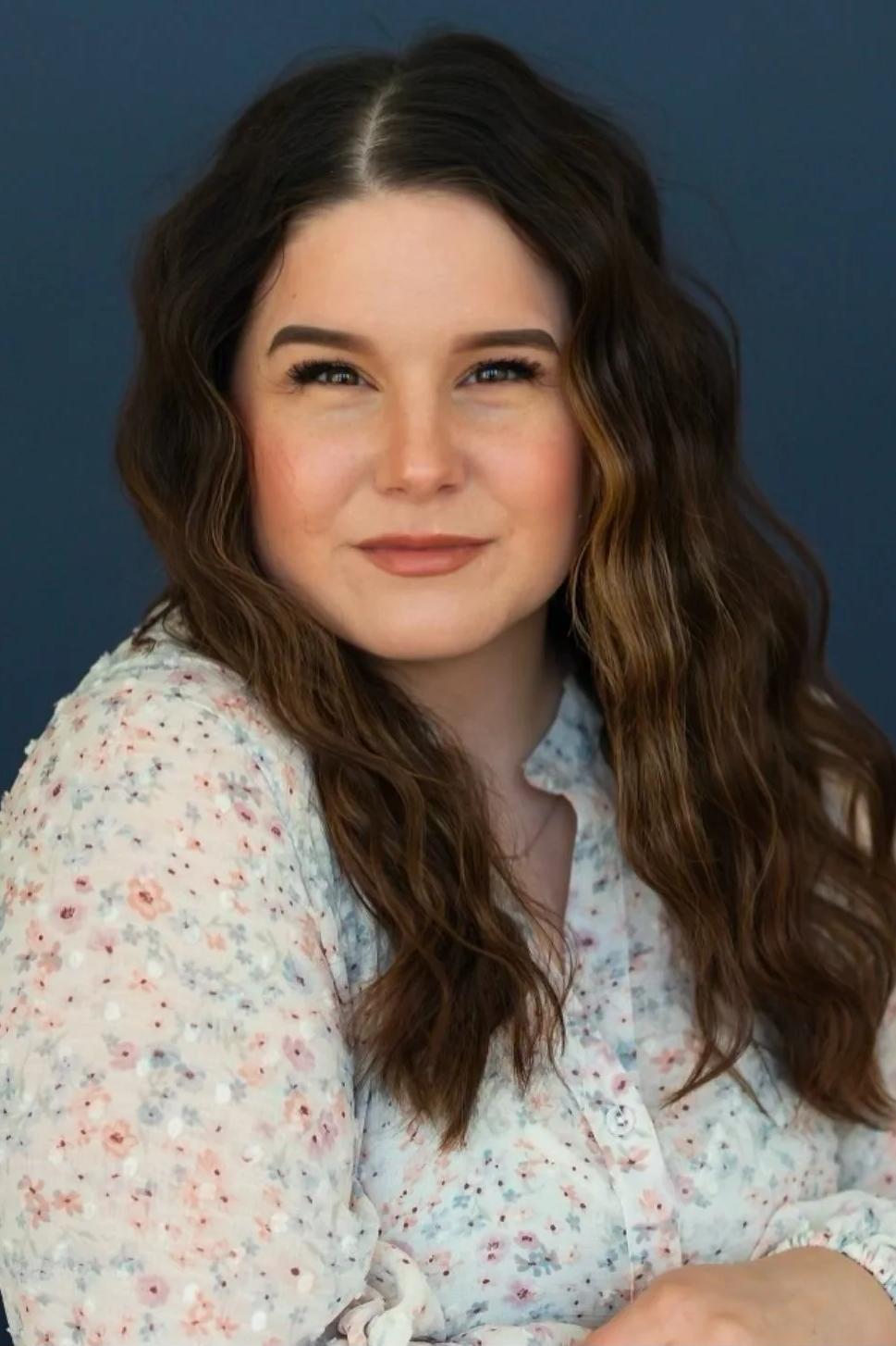 A young woman with long, wavy brown hair and a subtle smile, wearing a floral blouse, against a dark background.