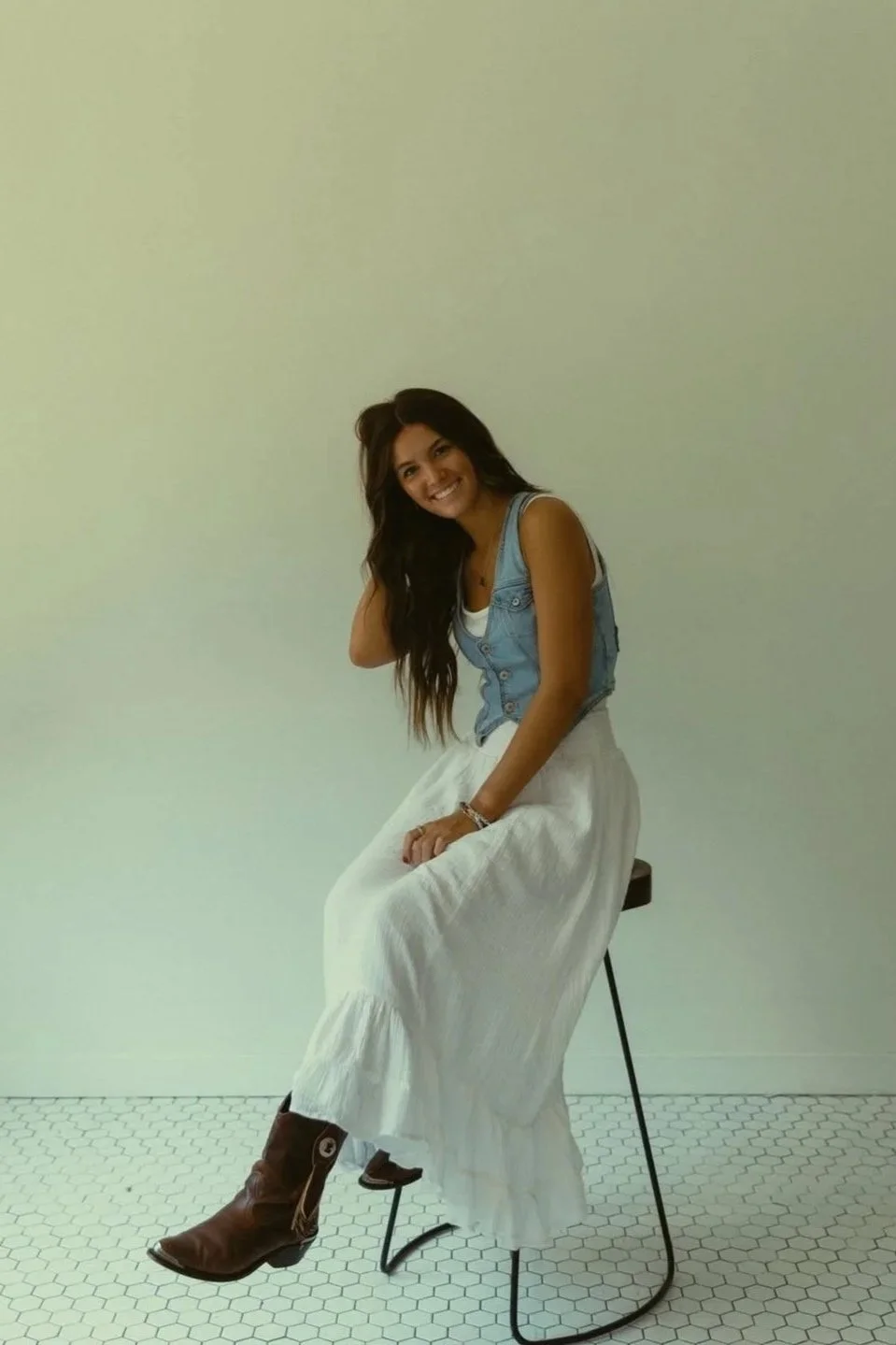 A young woman sitting on a stool, smiling, with long dark hair, wearing a sleeveless denim vest, white maxi skirt, and brown cowboy boots, in front of a plain off-white wall.
