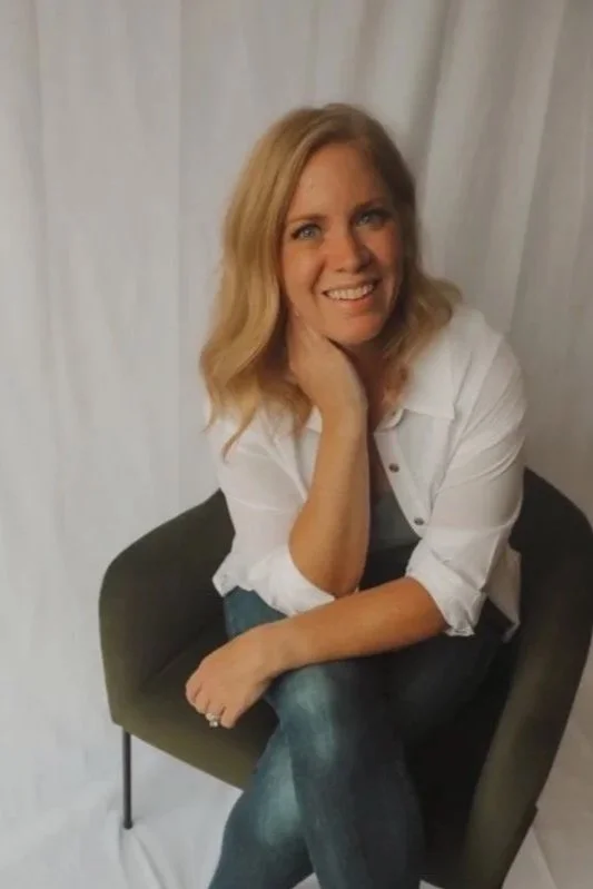 A woman with blonde hair, wearing a white shirt and jeans, sitting in a dark green chair with a light-colored curtain background, smiling and resting her head on her hand.