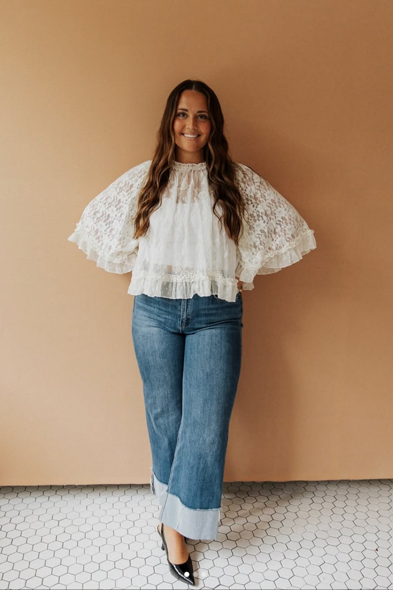 Woman with long wavy brown hair wearing a white lace blouse with puffy sleeves, jeans with wide cuffs, and black heels, standing against a beige wall.