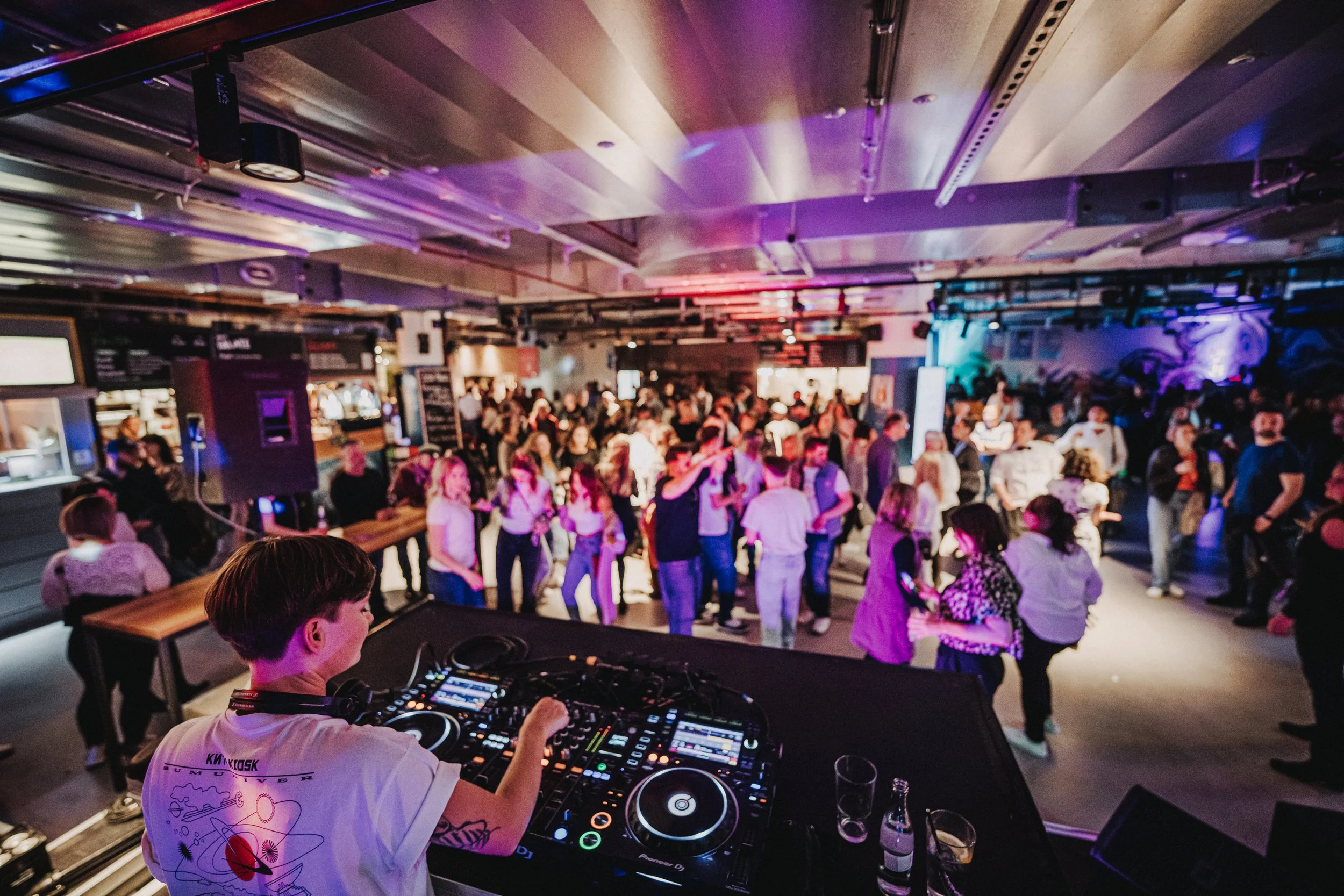 Menschen tanzen und feiern in einer Diskothek mit bunten Lichtern, vorne ein DJ am Mischpult, im Hintergrund eine tanzende Menge.