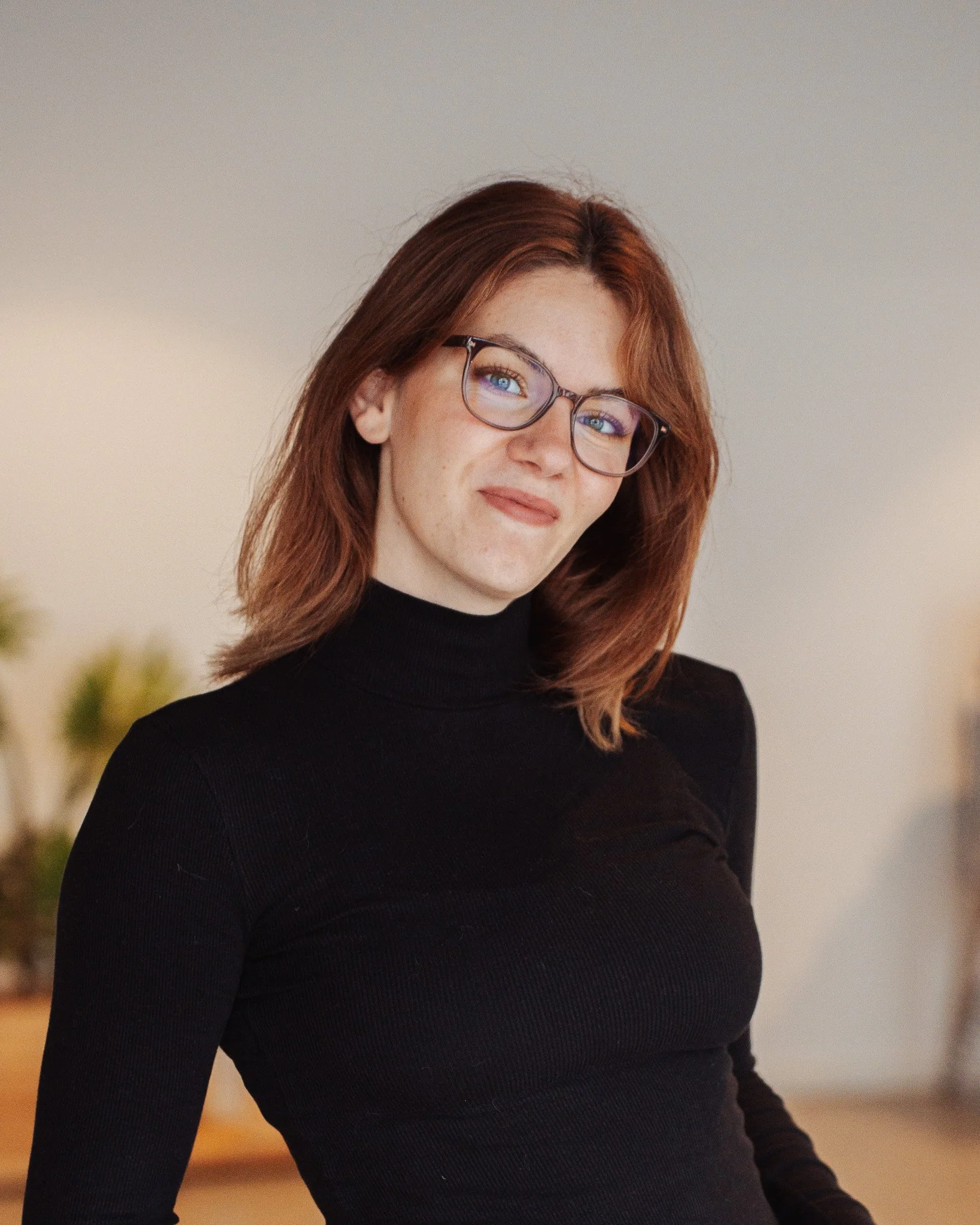Eine lachende Frau mit Brille, roten Haaren, weißen Hemd und Schmuck, im Hintergrund eine moderne Inneneinrichtung.