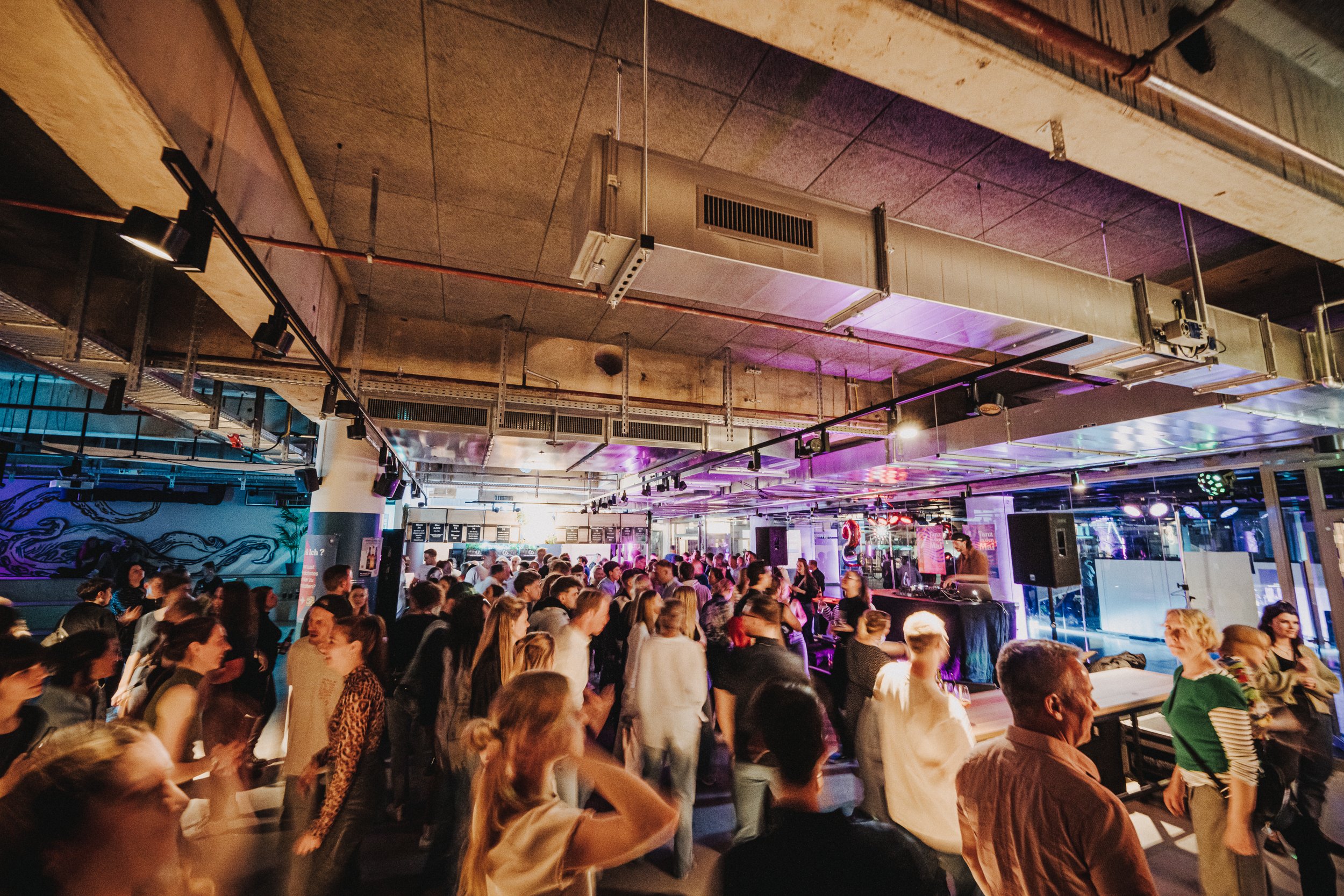 Ein Indoor-Event mit einer großen Menschenmenge, einer DJ-Person am DJ-Equipment auf der Bühne, bunte Lichter und eine lebhafte Atmosphäre.