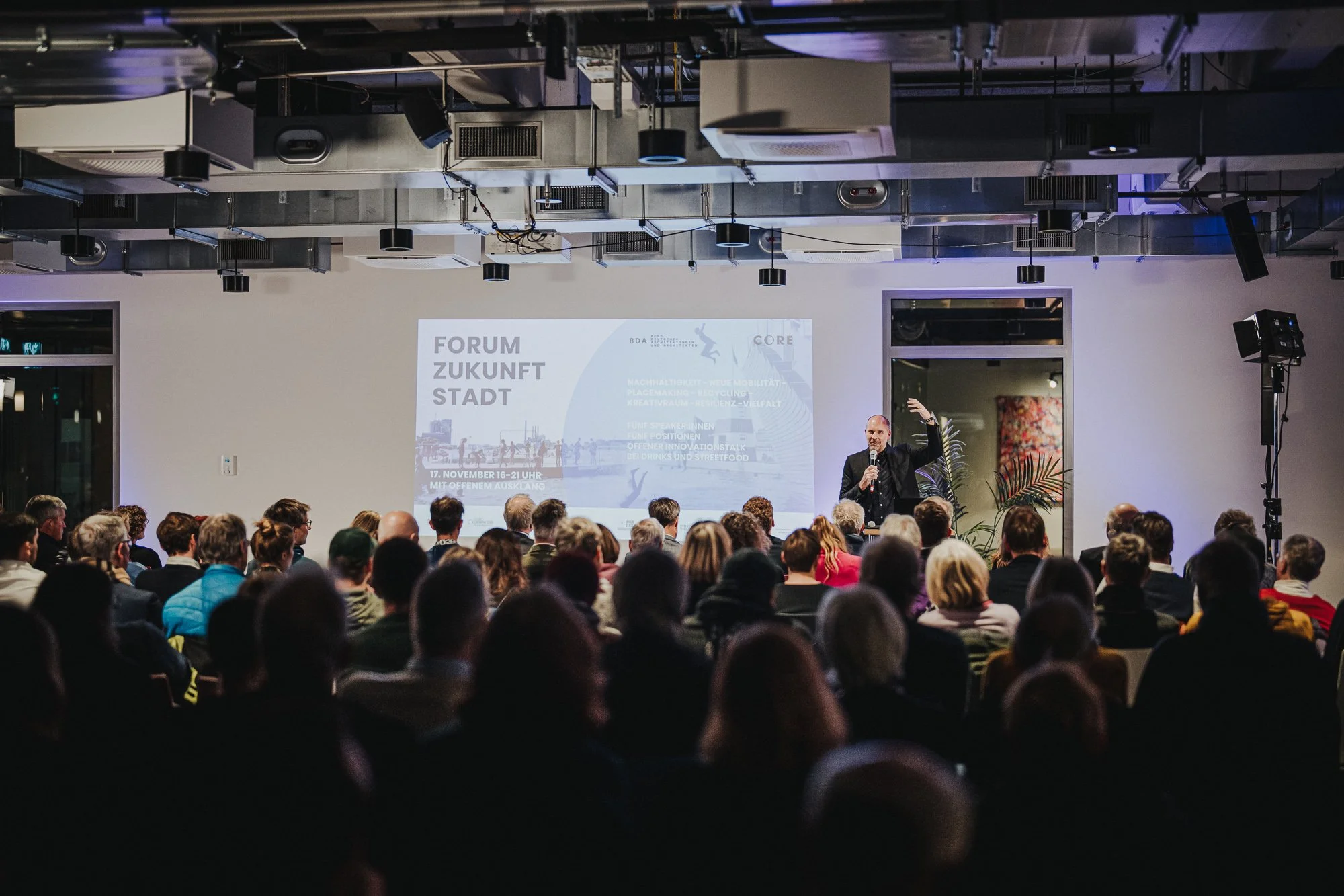 Ein Vortragender spricht vor einem Publikum in einem modernen Konferenzraum, während eine Präsentation an der Wand gezeigt wird.