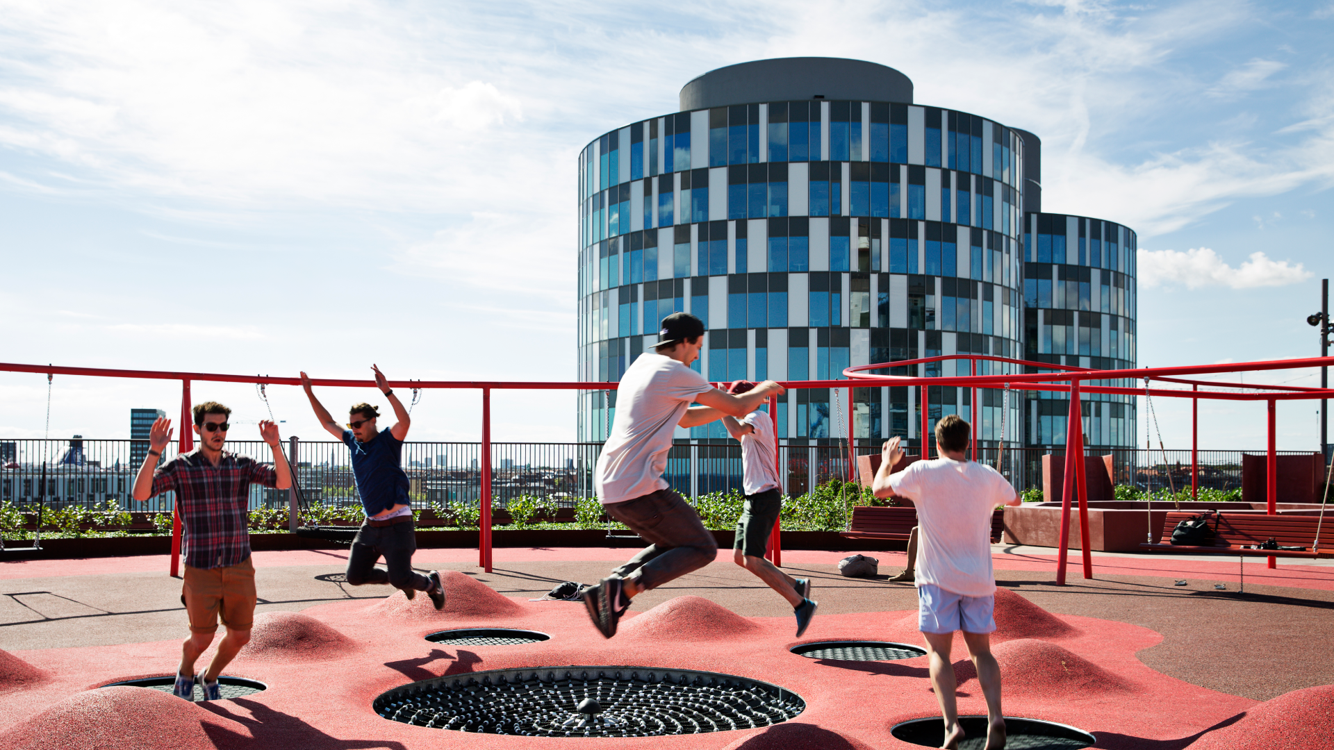 Jugendliche springen auf einem Trampolin auf einer Dachterrasse mit modernen Gebäuden im Hintergrund bei sonnigem Himmel.