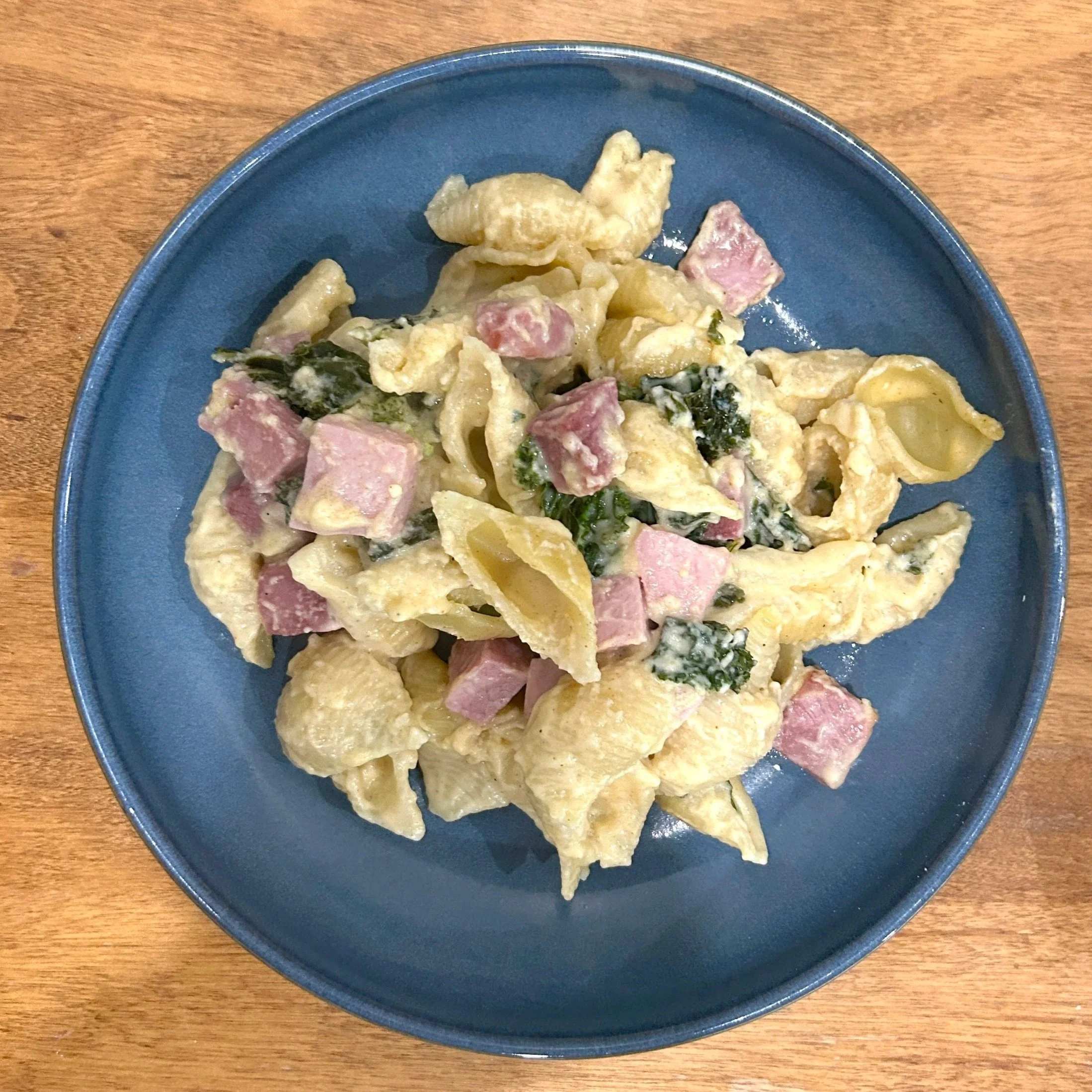 A bowl of pasta, ham and kale baked in a cheese sauce on a wooden table