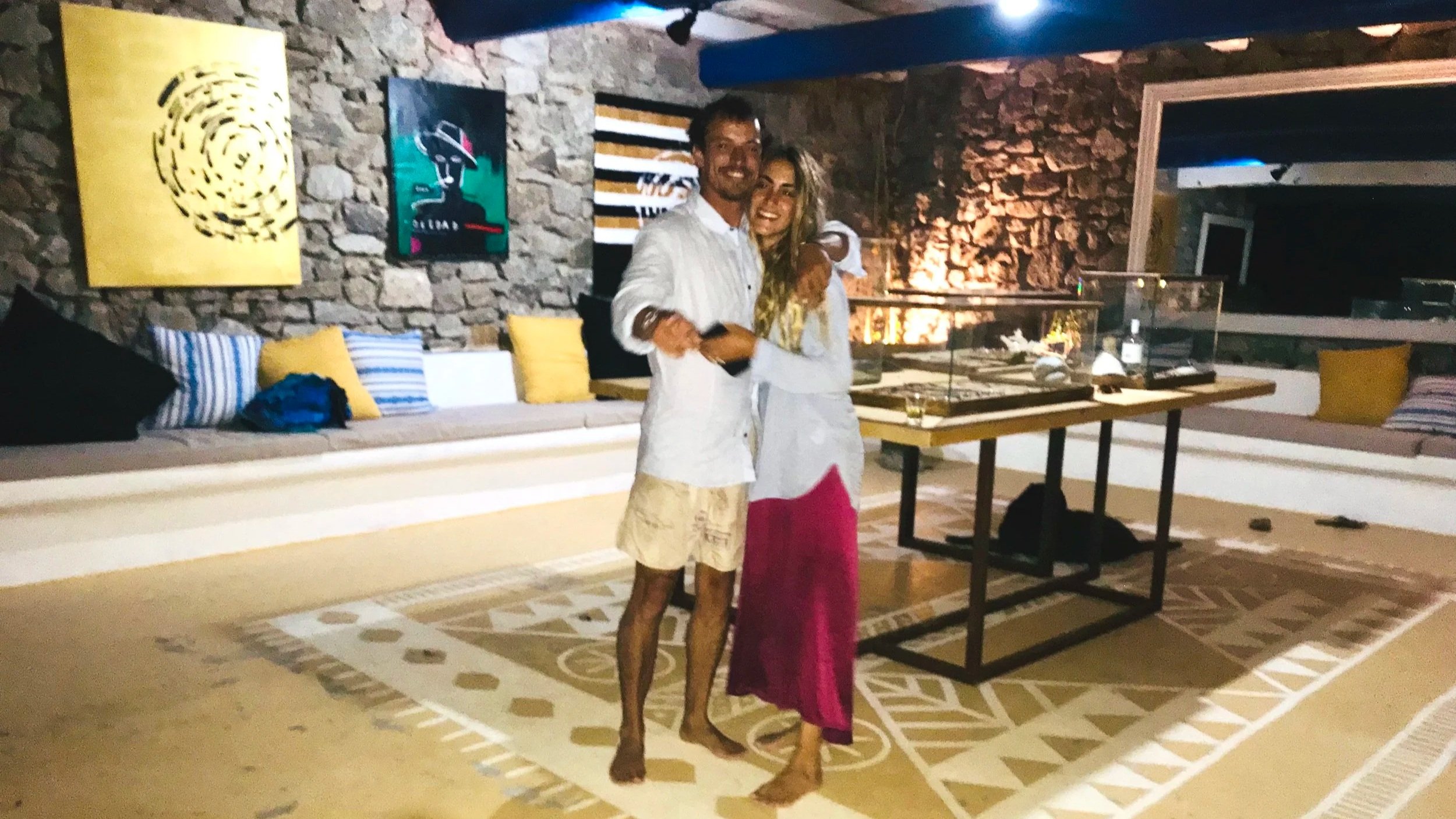 A man and woman hugging and smiling at a party or social gathering in a room with stone walls, colorful artwork, and a patterned rug. They appear to be holding a small black object, possibly a phone or camera.