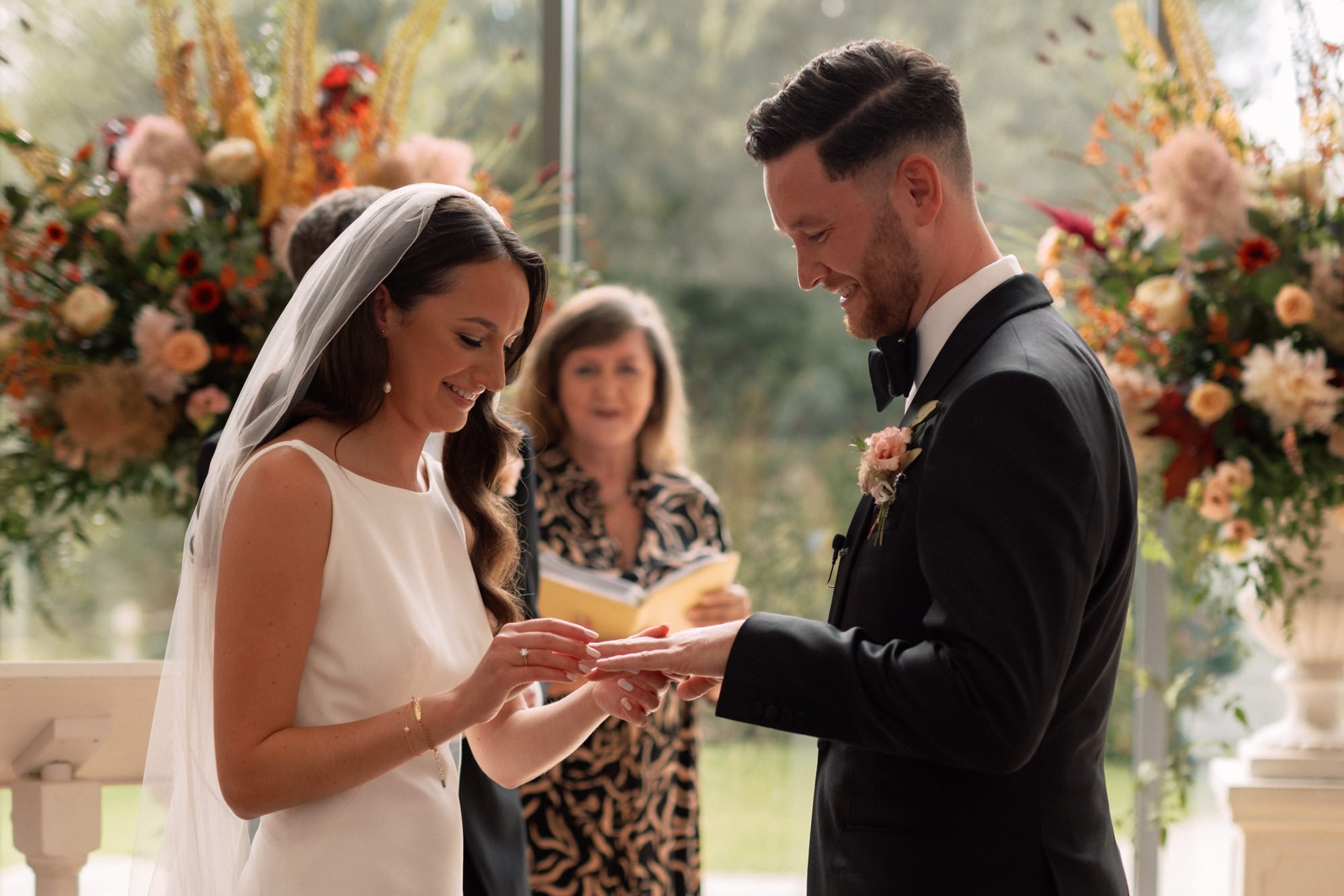 A bride places a wedding ring on her grooms fourth finger in their wedding ceremony at Millbridge Court, Farnham. Surrounded by beautiful flowers