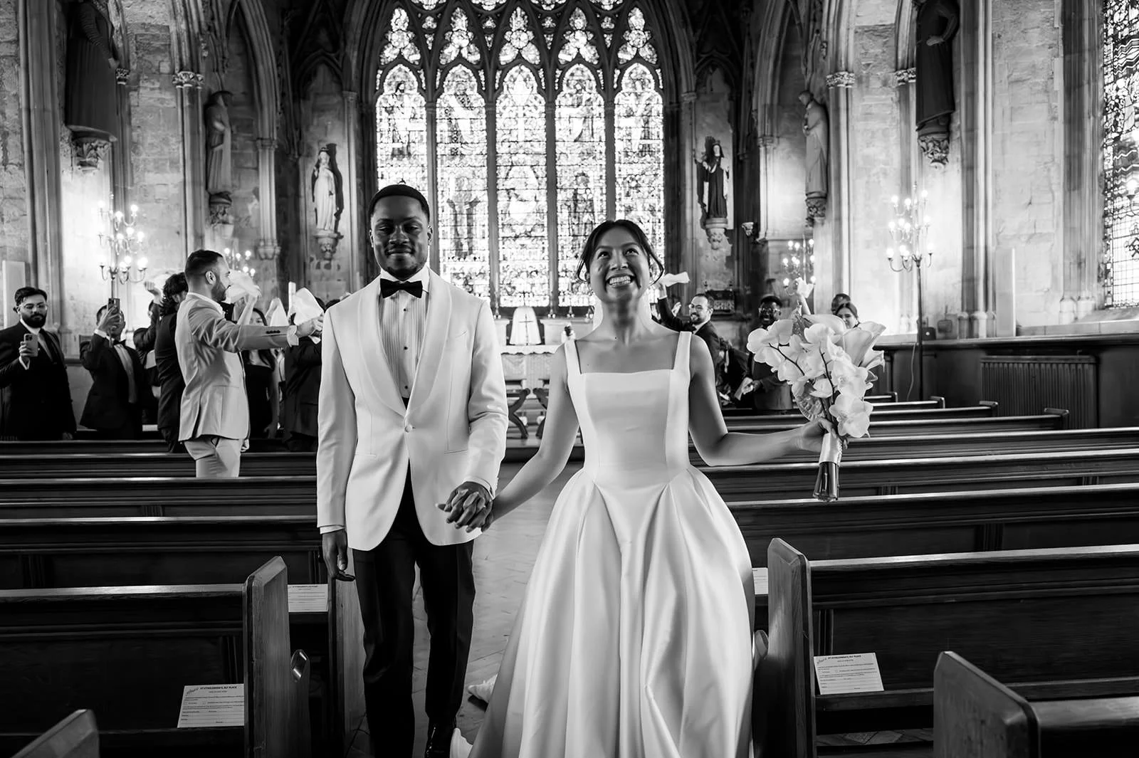 A newly married couple walking down the aisle of a church, holding hands. The bride is holding a bouquet and wearing a classic white wedding dress, while the groom is in a tuxedo with a bow tie. Guests are standing in the pews, taking photos and celebrating. The church has tall stained glass windows and ornate decorations.