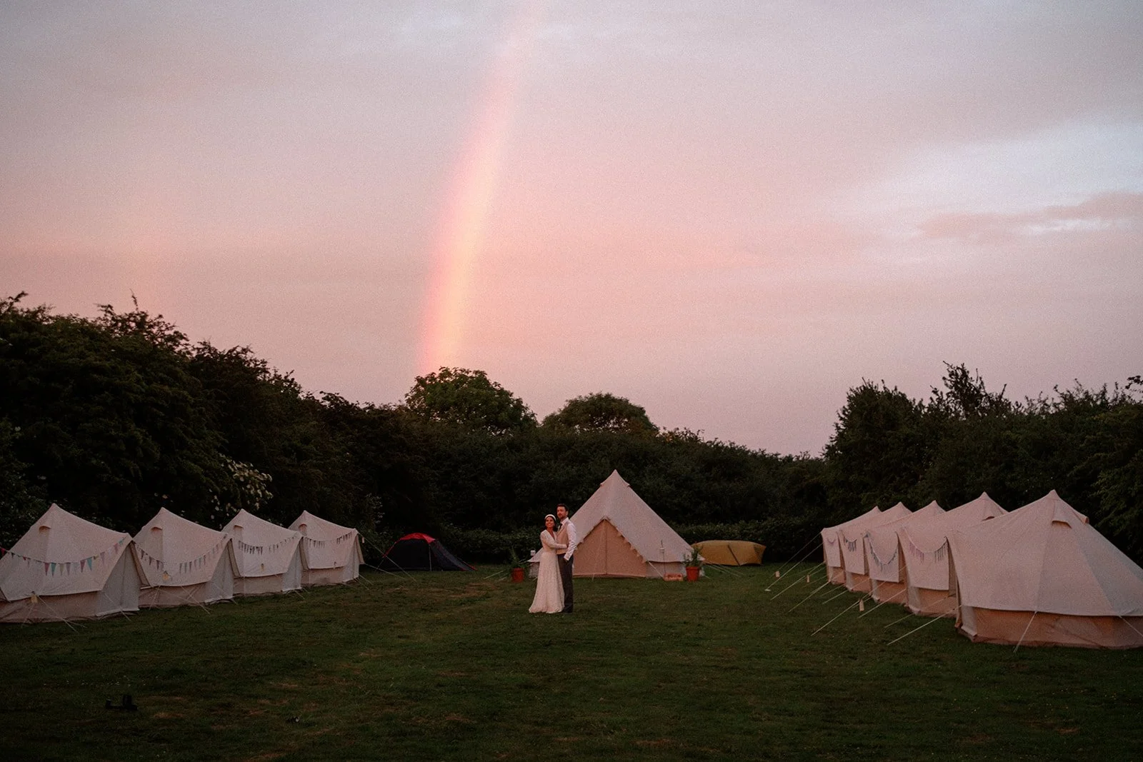 Newly weds enjoy a sunset rainbow amoungst tents they put out for their wedding guests in their garden on their wedding day.