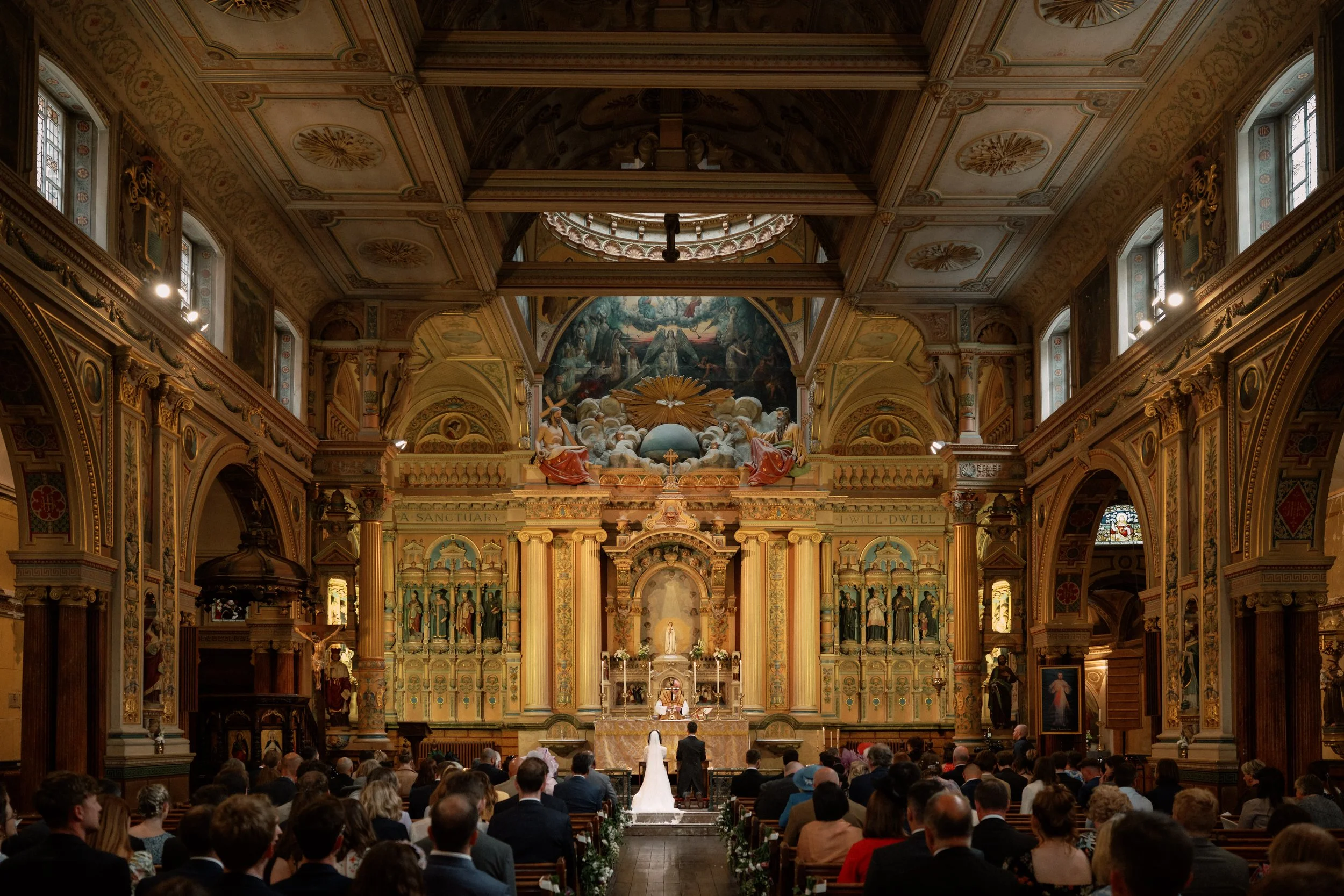 A wedding ceremony taking place inside a historic church with ornate gold and wood decor, a large congregation attending, a bride and groom at the altar, and religious artwork and statues adorning the walls.