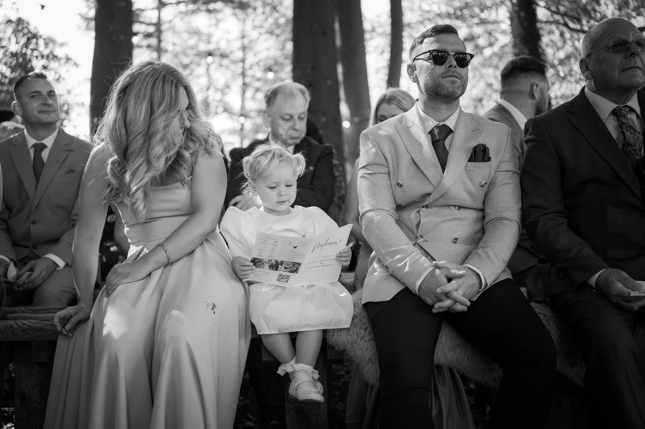 People sitting outdoors during a formal event, including a young girl reading a pamphlet, a woman in a gown, and men in suits, with trees in the background.