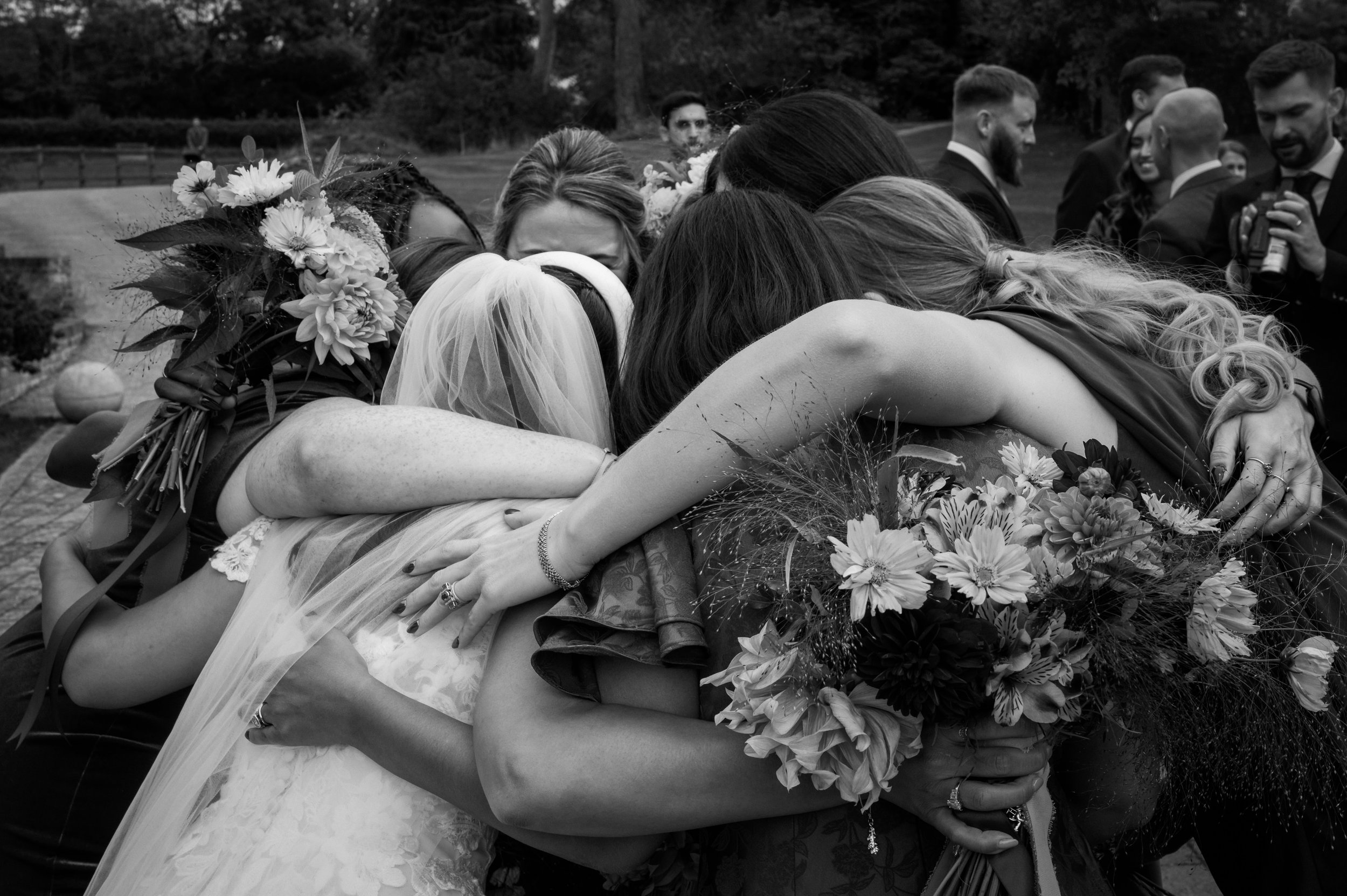 Group of women and men hugging at a wedding, with the bride in a wedding dress and veil holding a bouquet of flowers.
