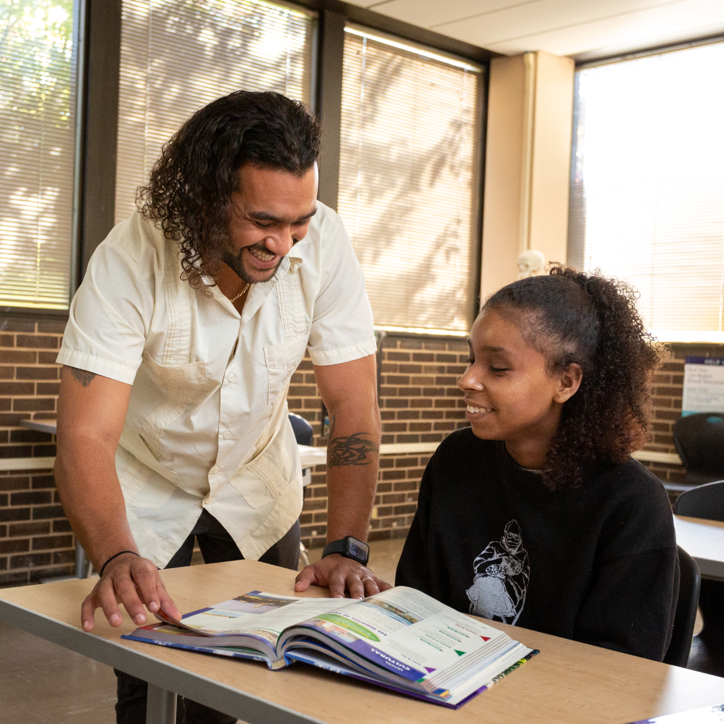 Teacher Carlos discusses text book with a middle school student in a classroom. The both have brown skin and long curly hair.