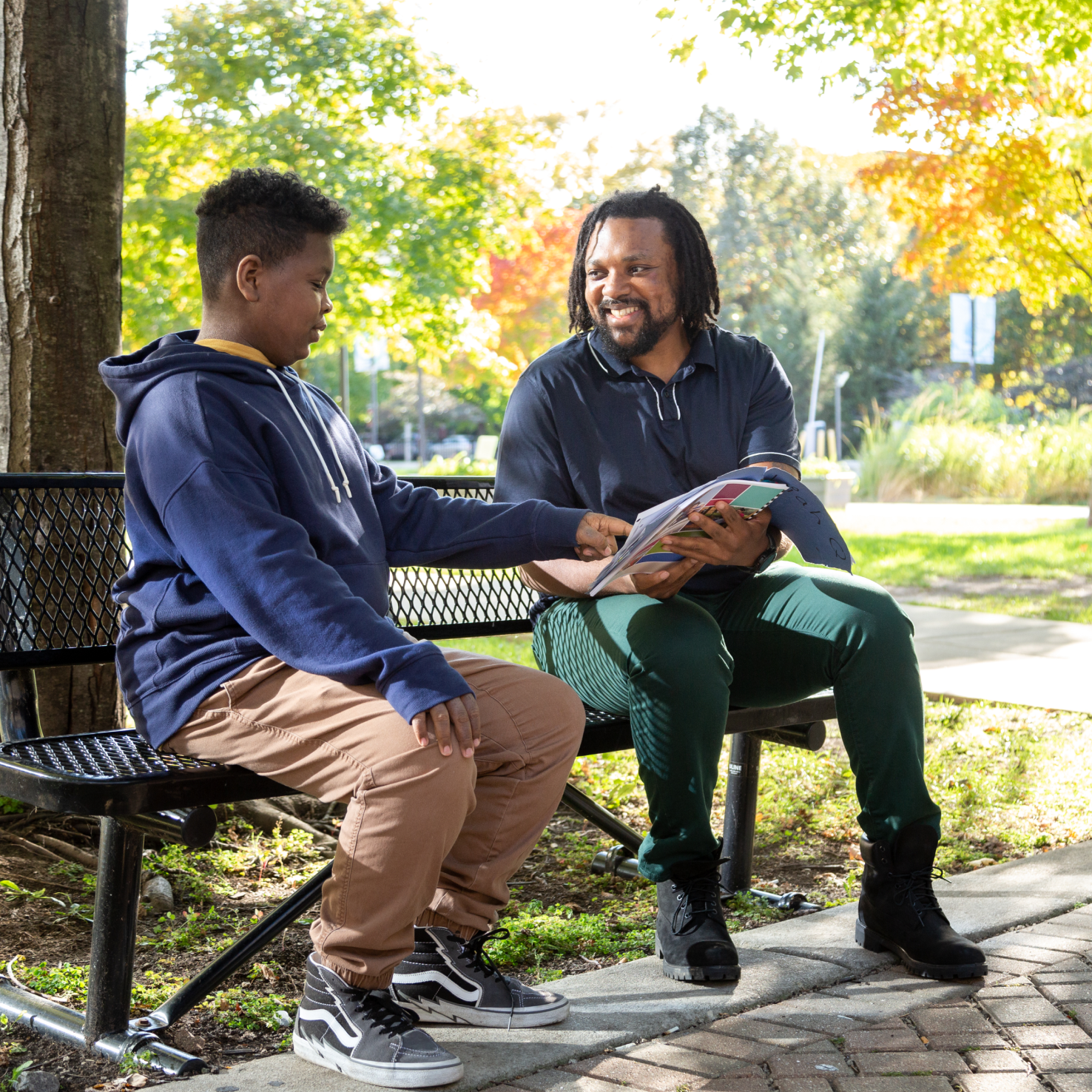 Teacher Andre and a young Black student sit on a park bench enjoying a book together.