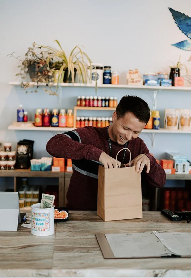 Product manager and budtender packing a paper bag at a store counter with shelves of products and plants in the background.