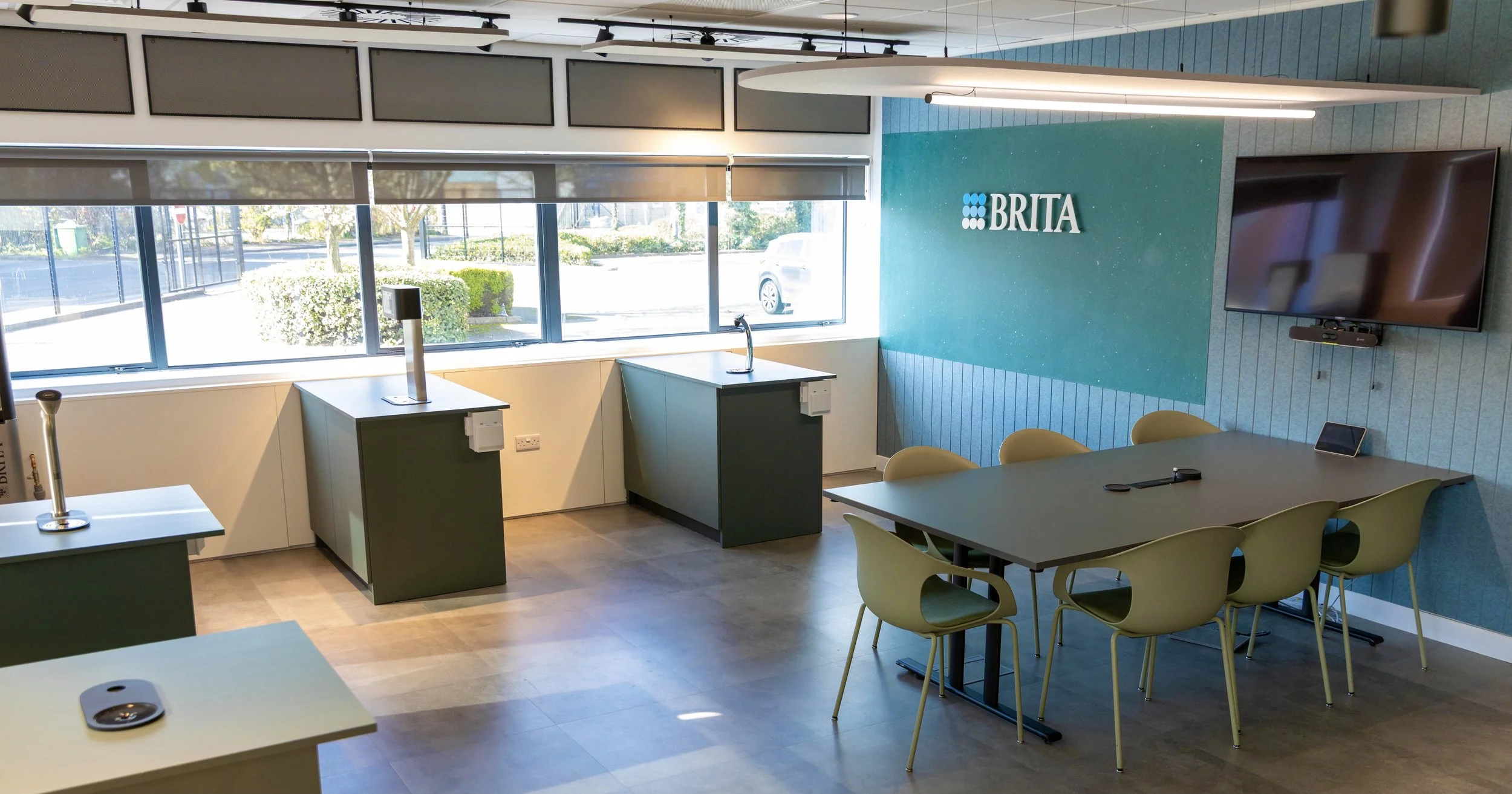 Interior of a training room a large table, yellow chairs, a wall-mounted TV, and a sign that says 'BRITA'. Contains two water dispensing stations near the window, which has blinds and a view of trees and parked cars outside.