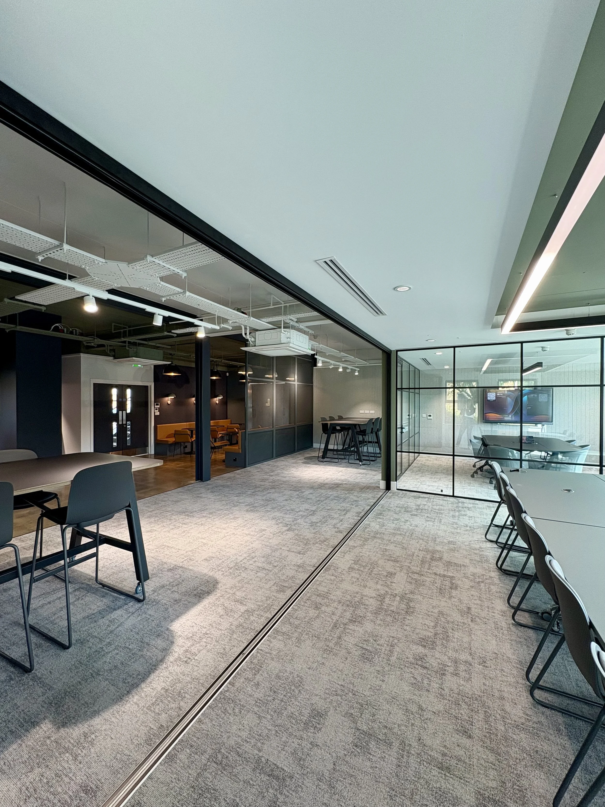 Modern office space with glass-walled conference meeting room, tables, chairs, and ceiling lighting fixtures.