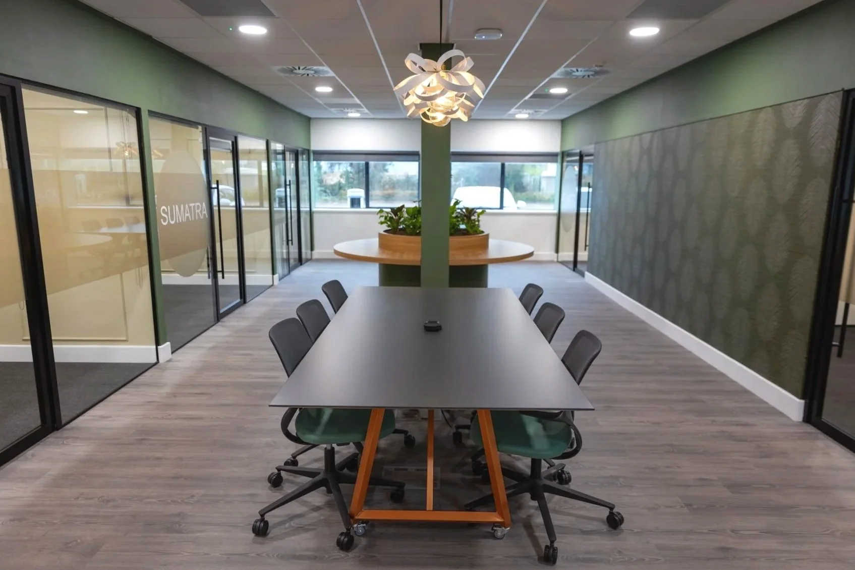 Modern office with a decorative chandelier hanging from the ceiling, a green painted wall, and glass partition walls with frosted sections on both sides; a central pillar with a circular planter and green plants, and windows allowing natural light.