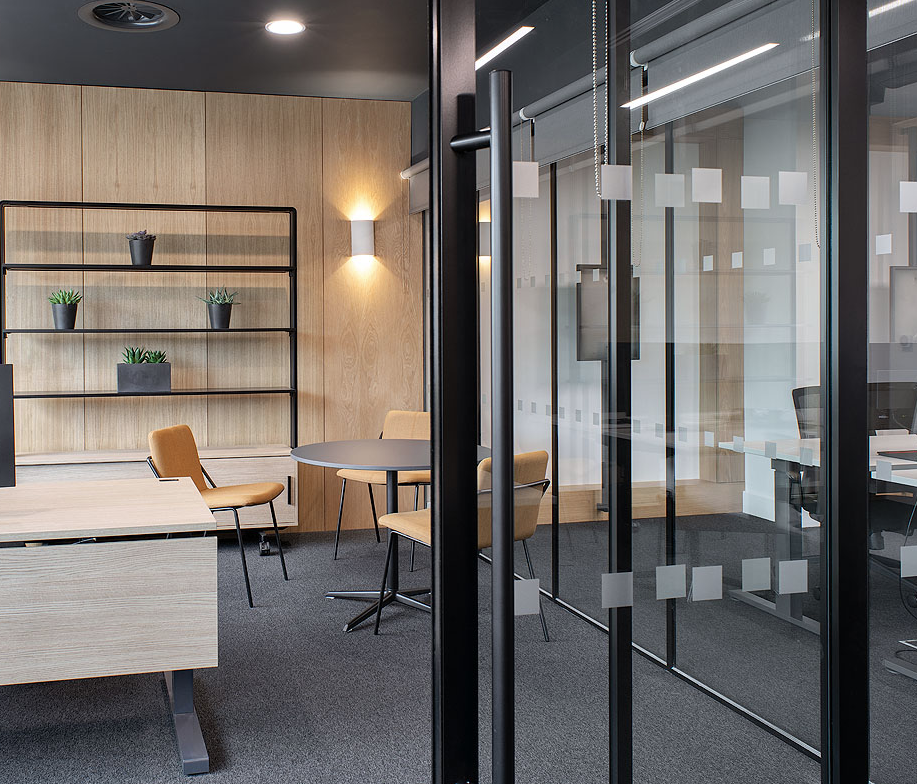 Modern office meeting room with glass walls, beige chairs, round table, and wall-mounted shelves with potted plants.