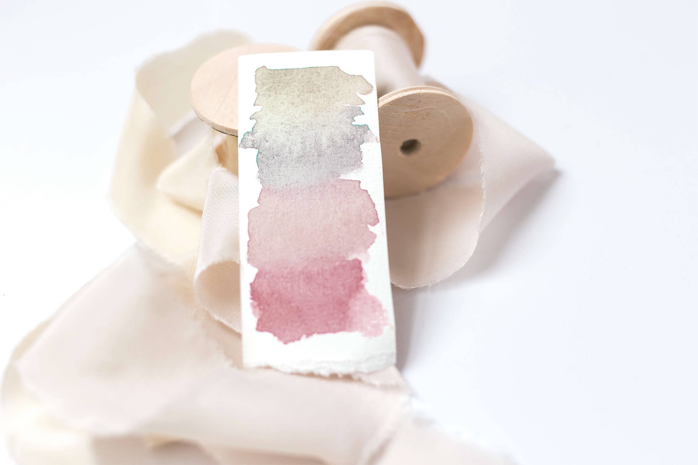 Watercolor swatch palette with shades of gray, pink, and purple, resting on a white cloth with wooden spools in the background.