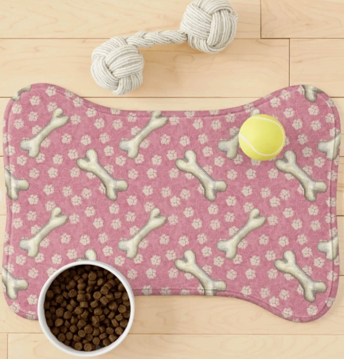 A pink dog feeding mat with bone and flower pattern, a yellow tennis ball, a white bowl filled with dry dog food, and a knotted rope toy on a wooden floor.