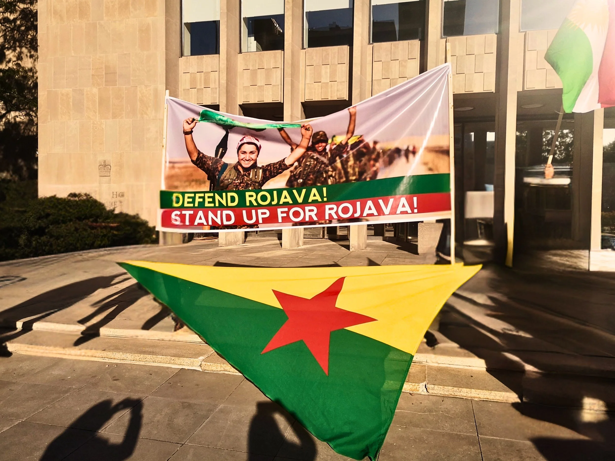 A banner with Kurdish resistance fighters holding the peace sign with the slogans "Defend Rojava and Stand Up for Rojava" written on the bottom. A flag of the Kurdish resistance movement in the foreground.