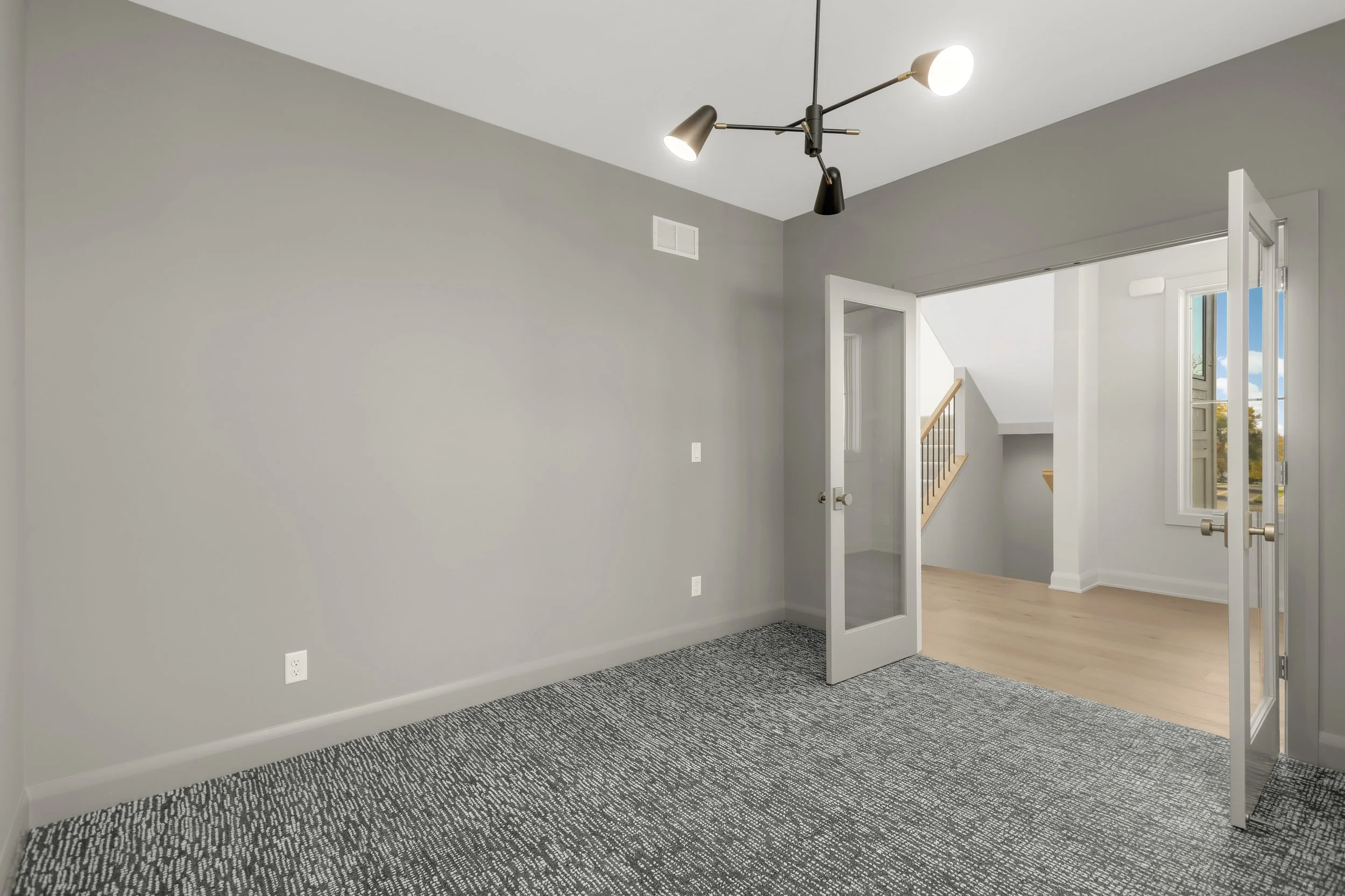Modern carpeted room with gray walls, black ceiling light fixture, glass-paneled double doors, and view of a staircase and foyer.