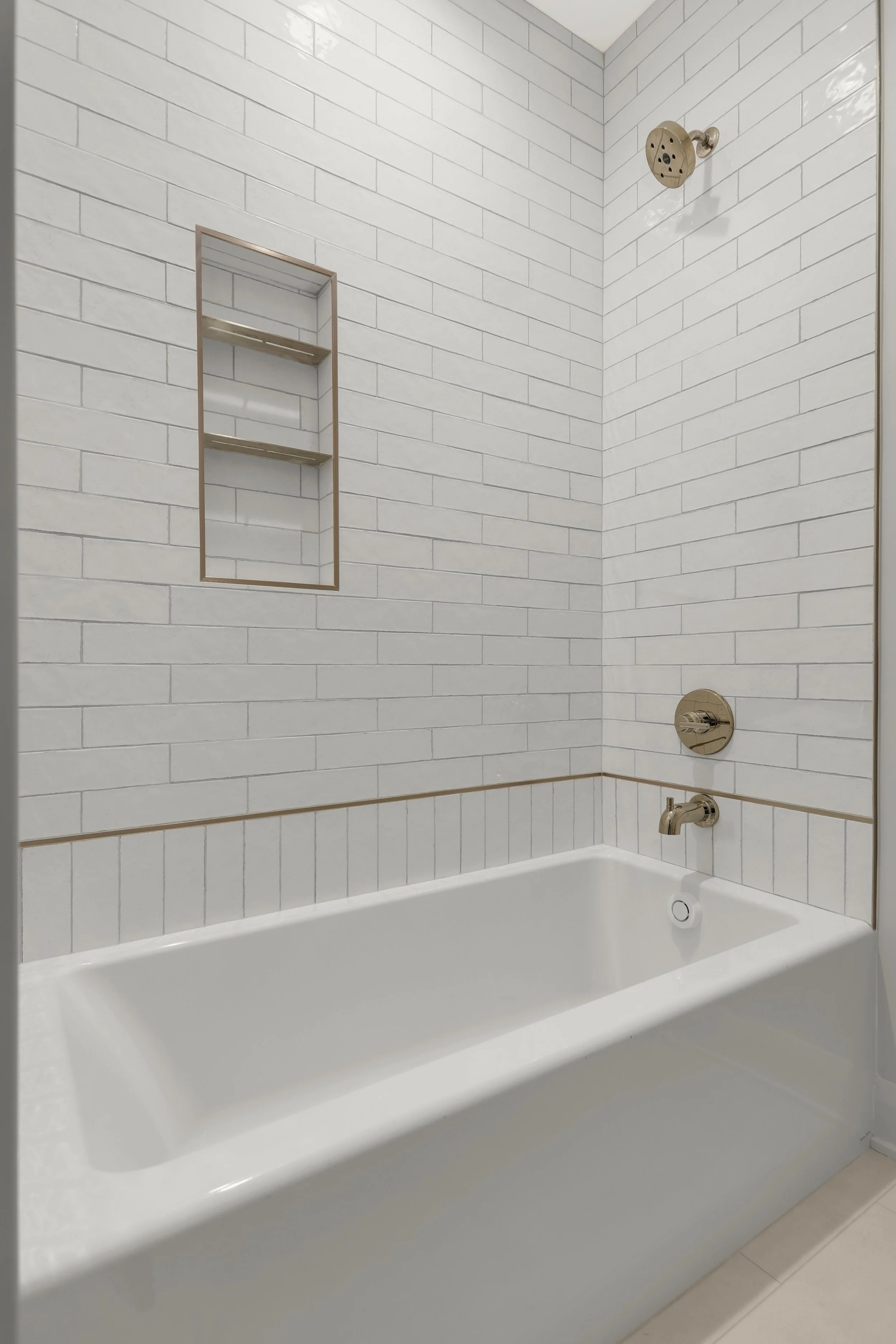 White subway-tile bathroom with a bathtub, brushed brass fixtures, showerhead, recessed shelf, and wall-mounted faucet.