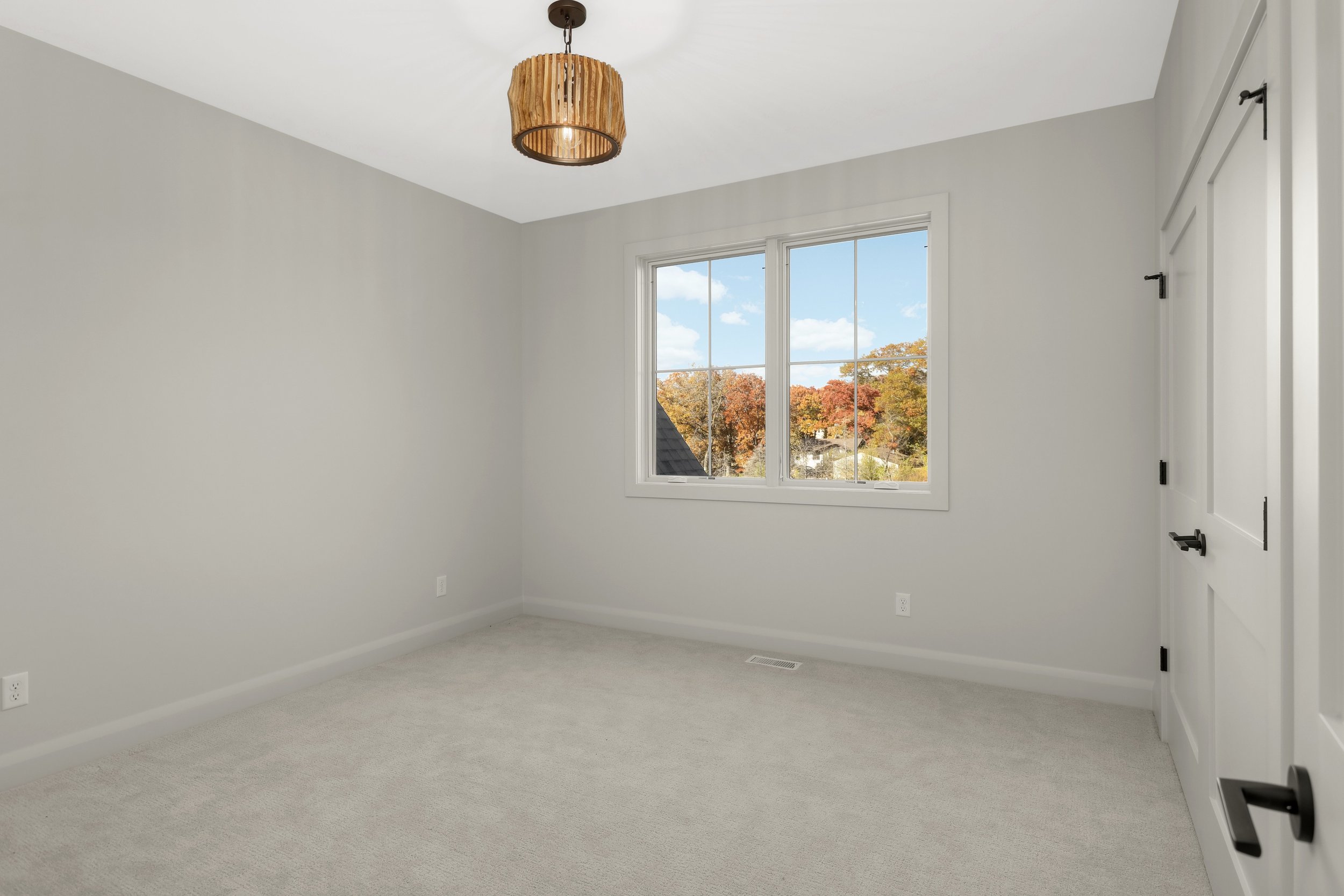 Empty carpeted bedroom with white walls, a wooden pendant light, double window, and white paneled door overlooking autumn trees.