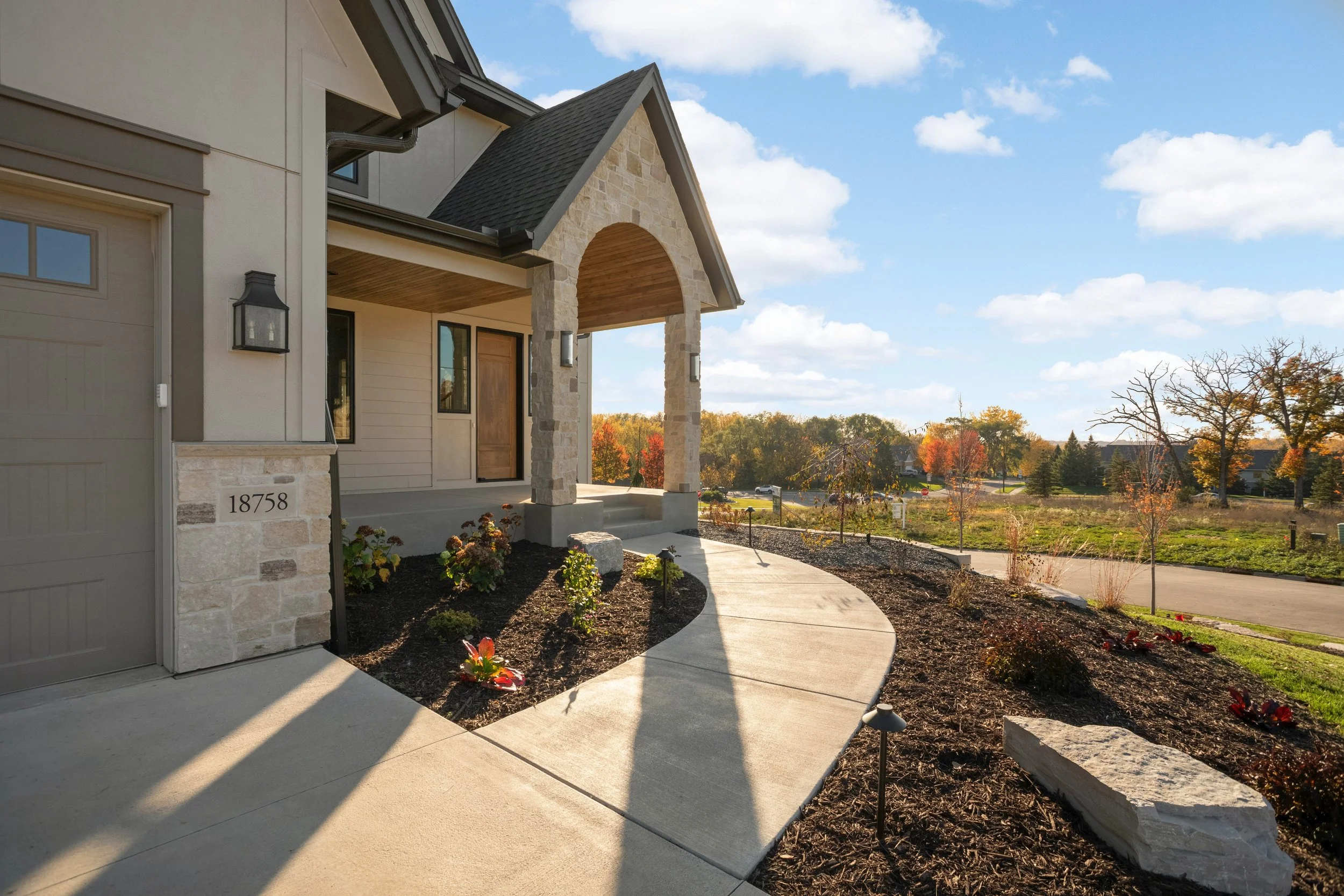 Stone-accented suburban home entrance with covered porch, landscaped walkway, garage, and autumn neighborhood view