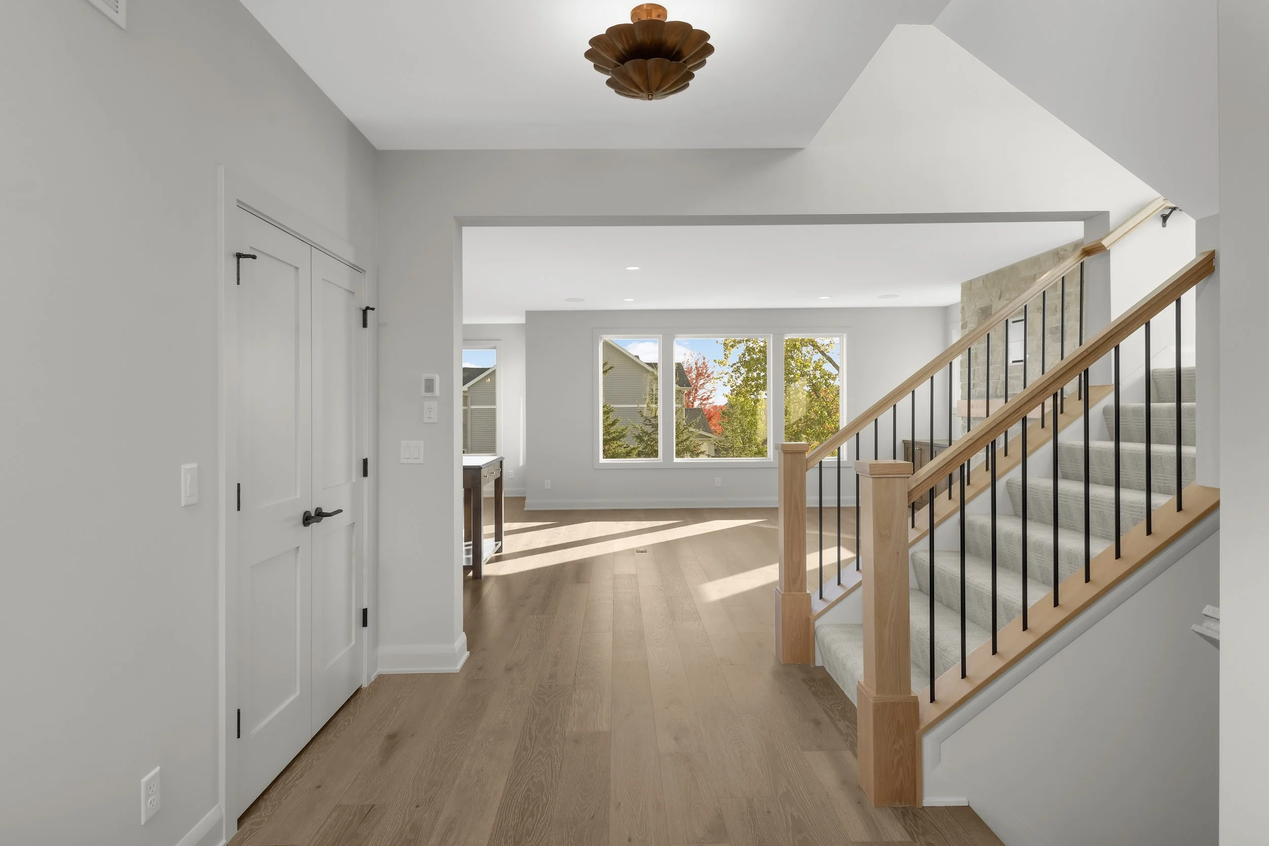 Bright modern home entryway with hardwood floors, carpeted staircase, black metal railing, white doors, and large windows.