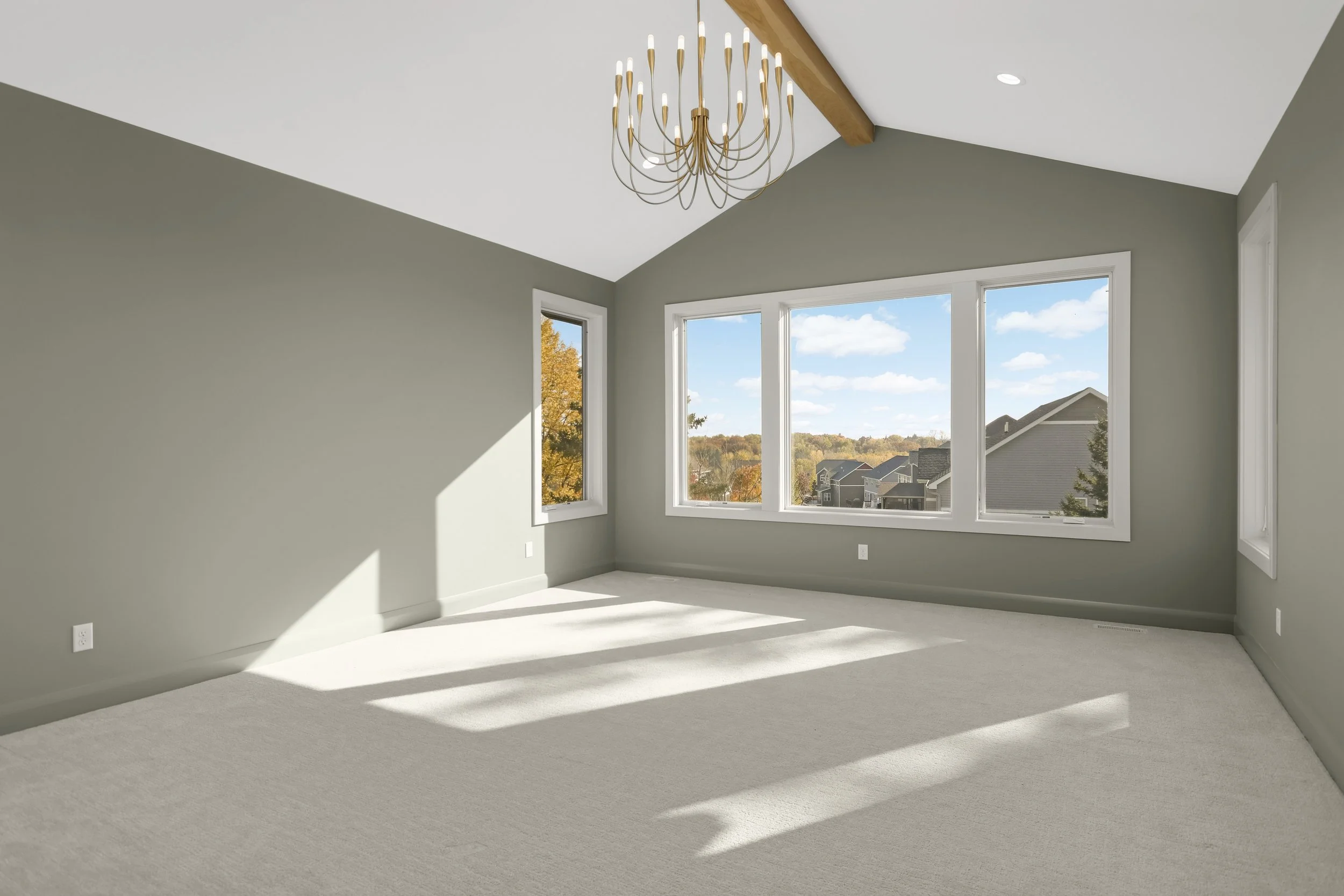 Empty sunlit room with gray walls, carpeted floor, large windows overlooking suburban homes, and a modern gold chandelier