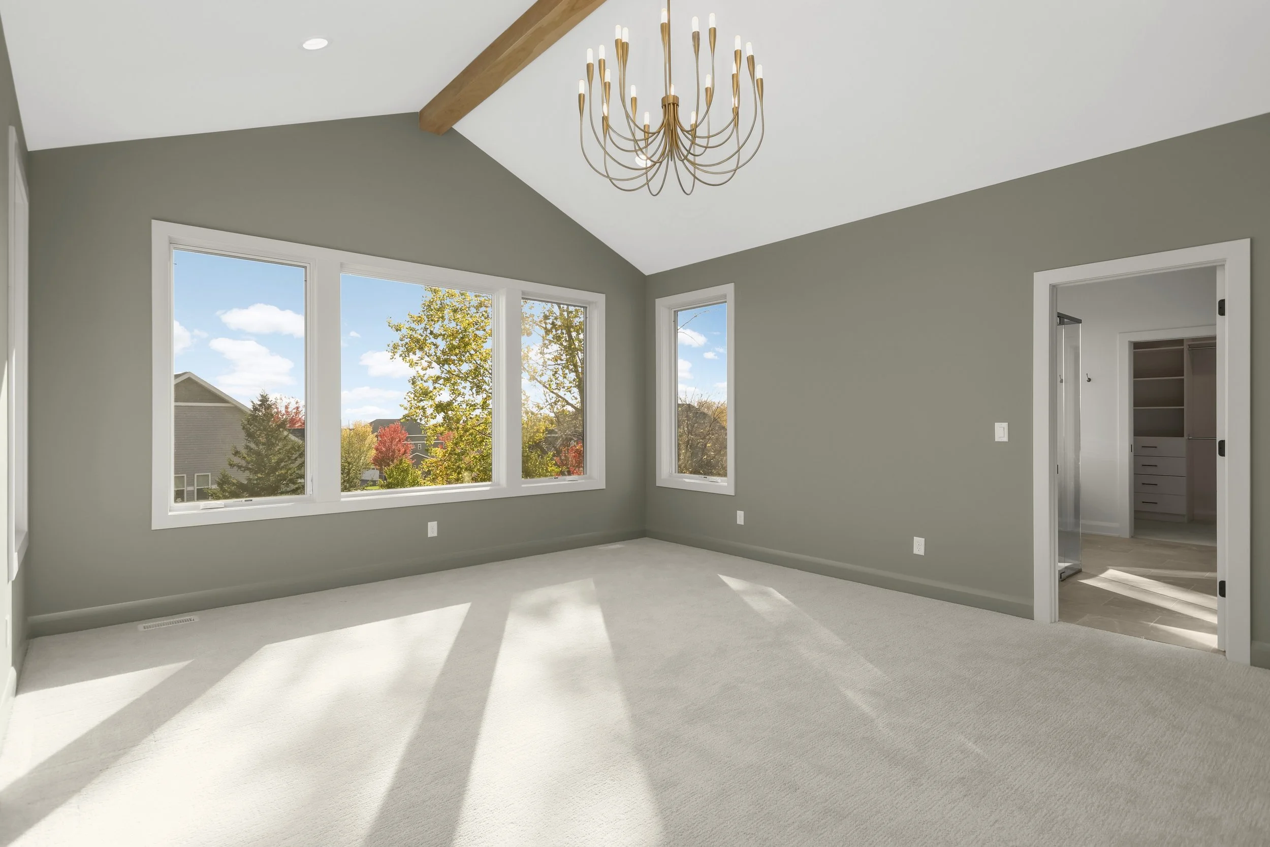Bright empty bedroom with gray walls, white carpet, large windows, exposed wood ceiling beam, gold chandelier, and walk-in closet.