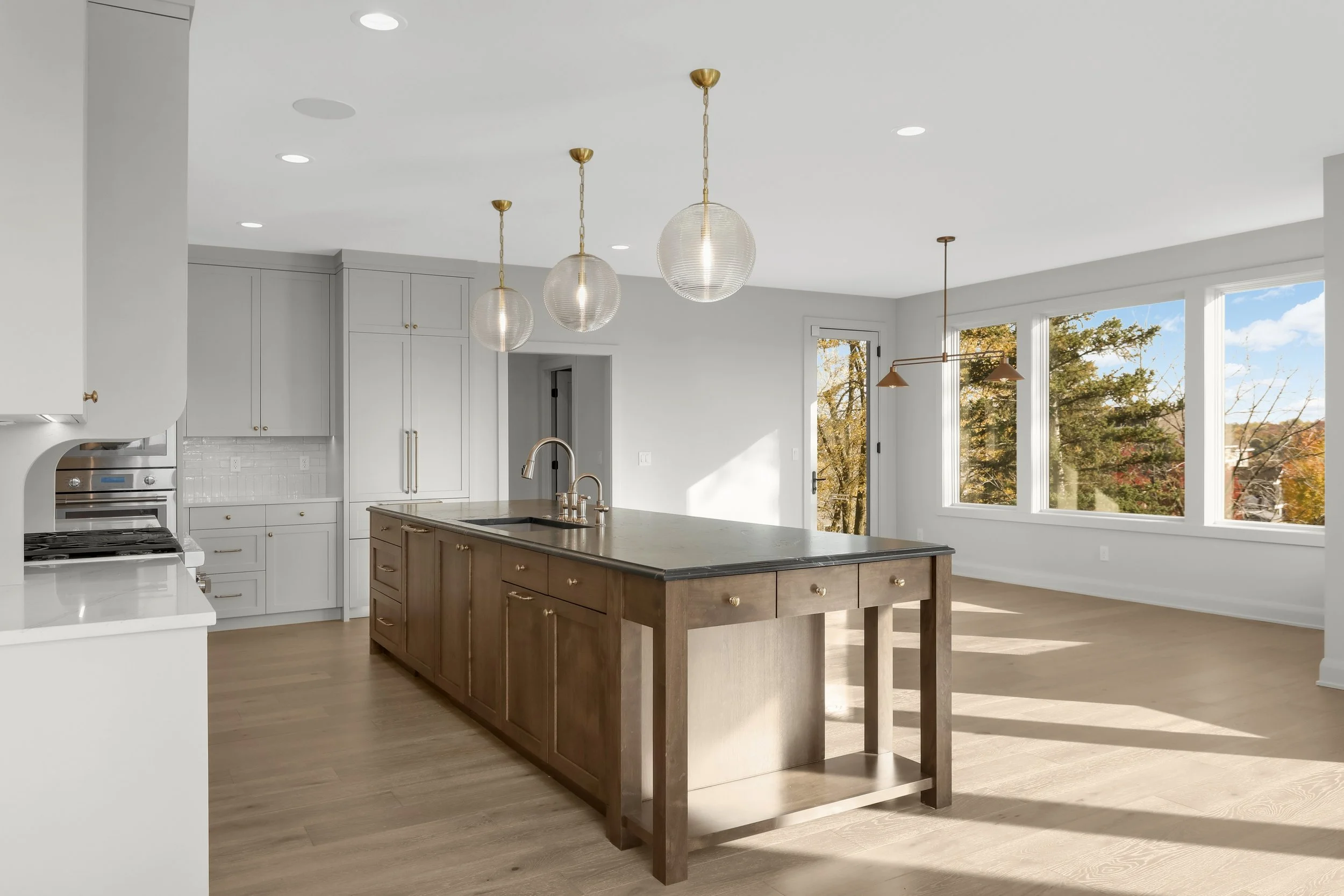 Bright modern kitchen with a large wood island, white cabinets, globe pendant lights, stainless steel appliances, and large windows overlooking trees