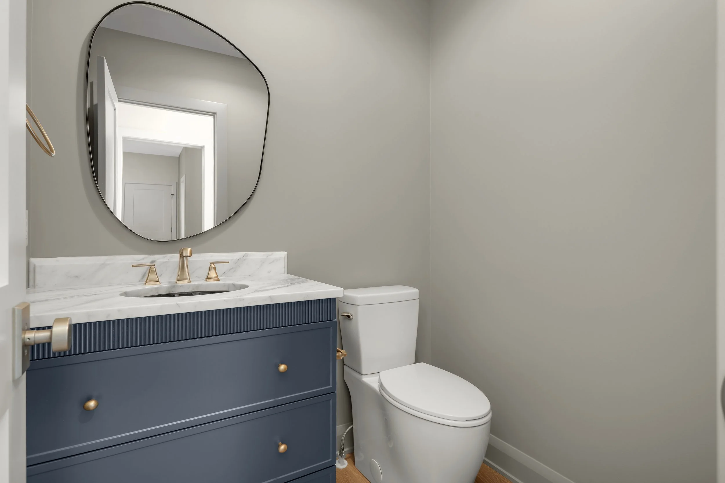 Modern bathroom with navy blue vanity, white marble countertop, gold fixtures, rounded mirror, and white toilet