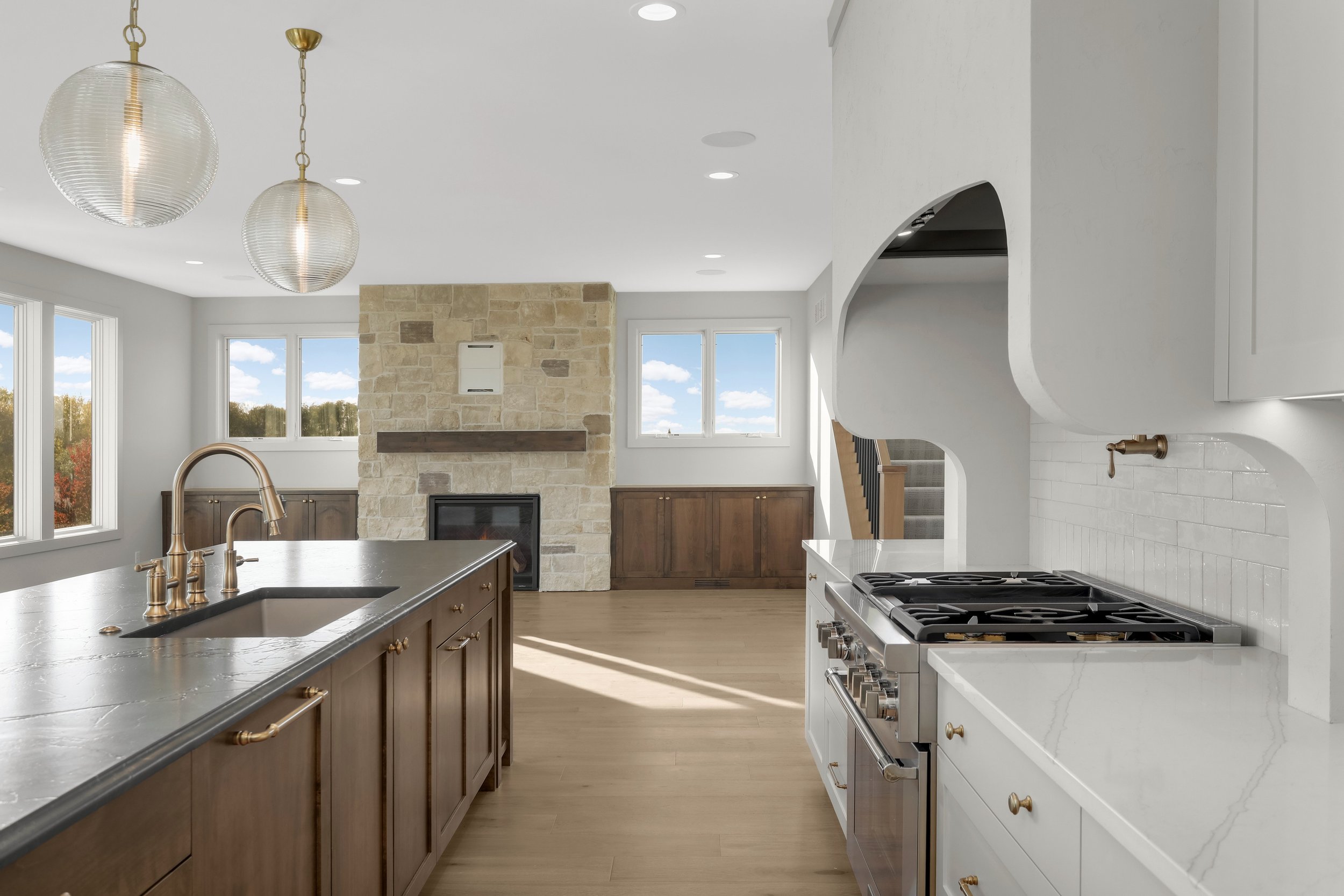 Bright modern kitchen with a large island, brass fixtures, pendant lights, stone fireplace, white cabinetry, and stainless steel range.