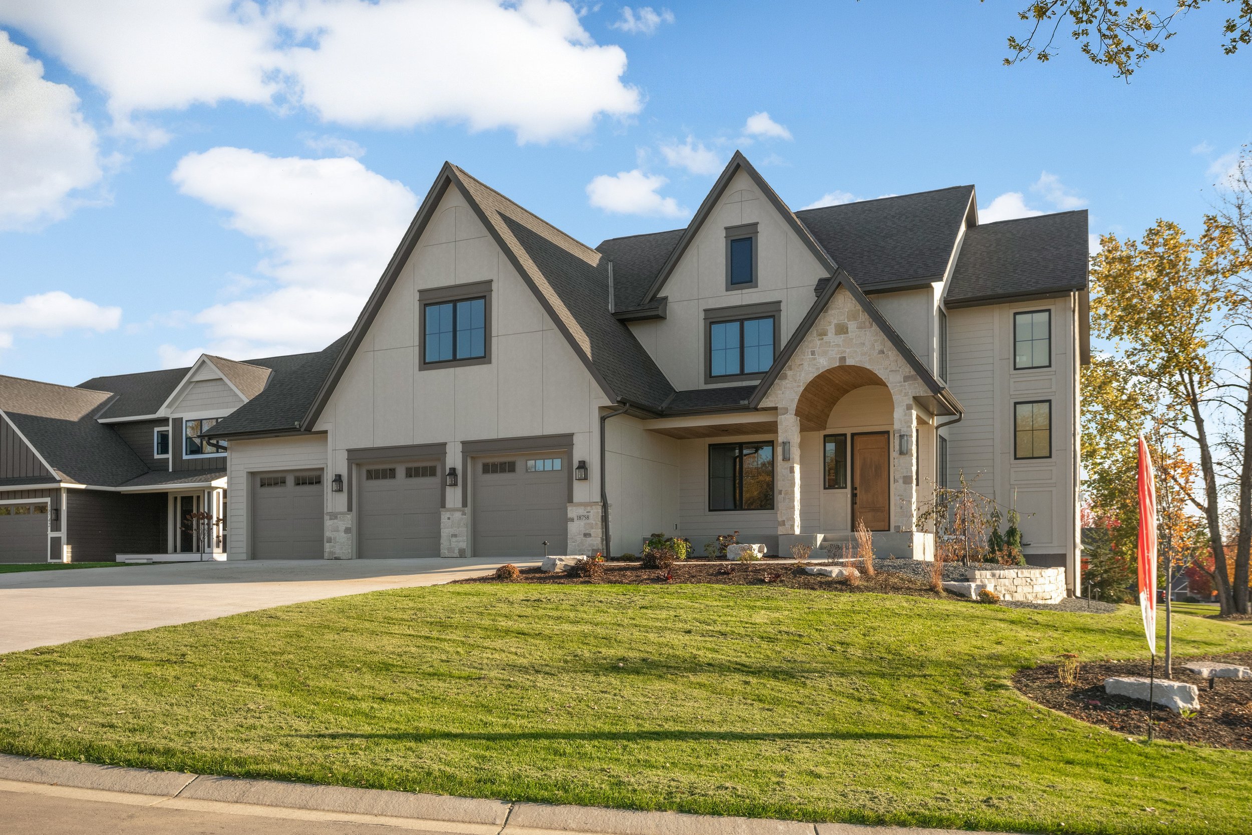 Large modern suburban home with multiple gabled roofs, stone entryway, three-car garage, landscaped yard, and neighboring houses