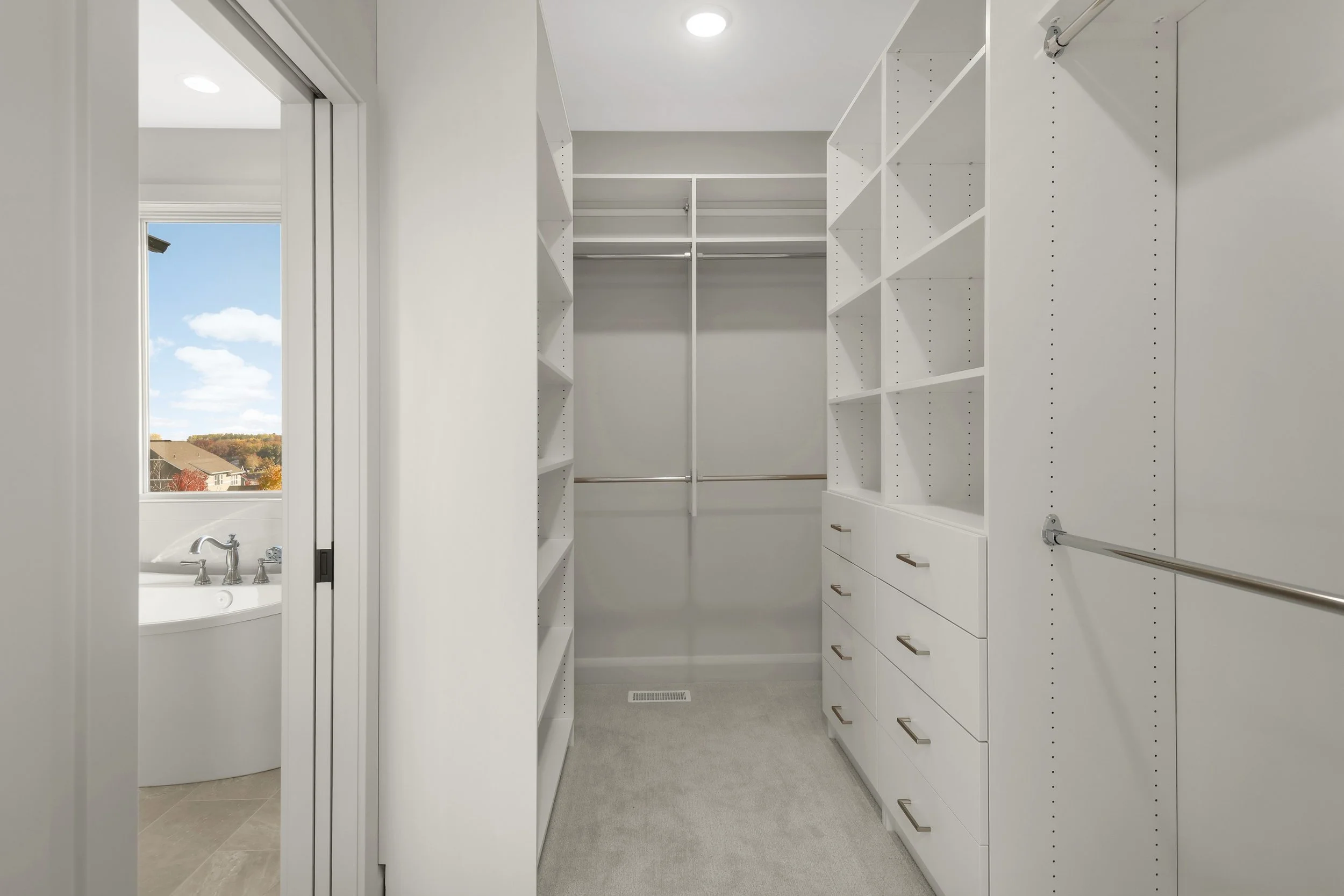 White walk-in closet with built-in shelving, drawers, hanging rods, and a bathroom visible through the doorway