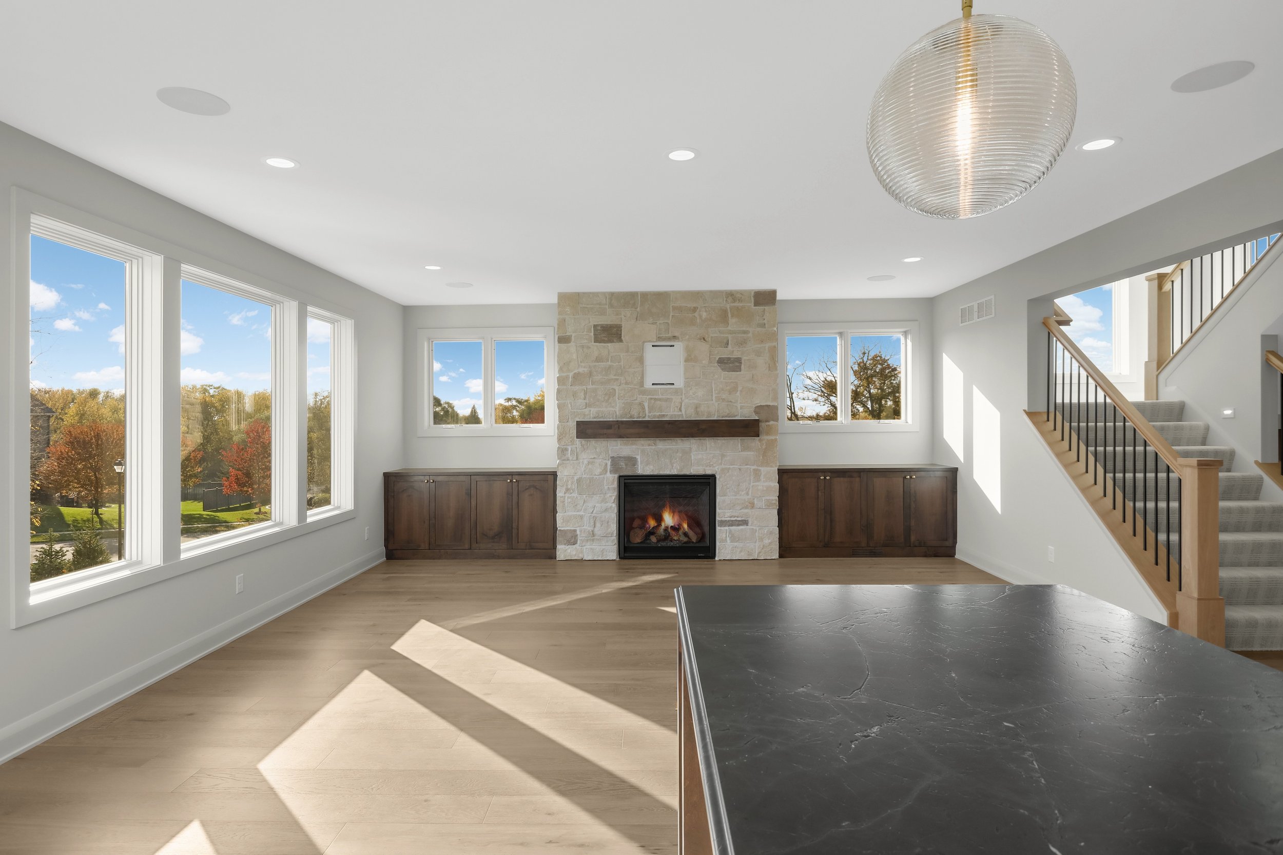 Bright modern living room with a stone fireplace, built-in cabinets, large windows, staircase, and black kitchen island