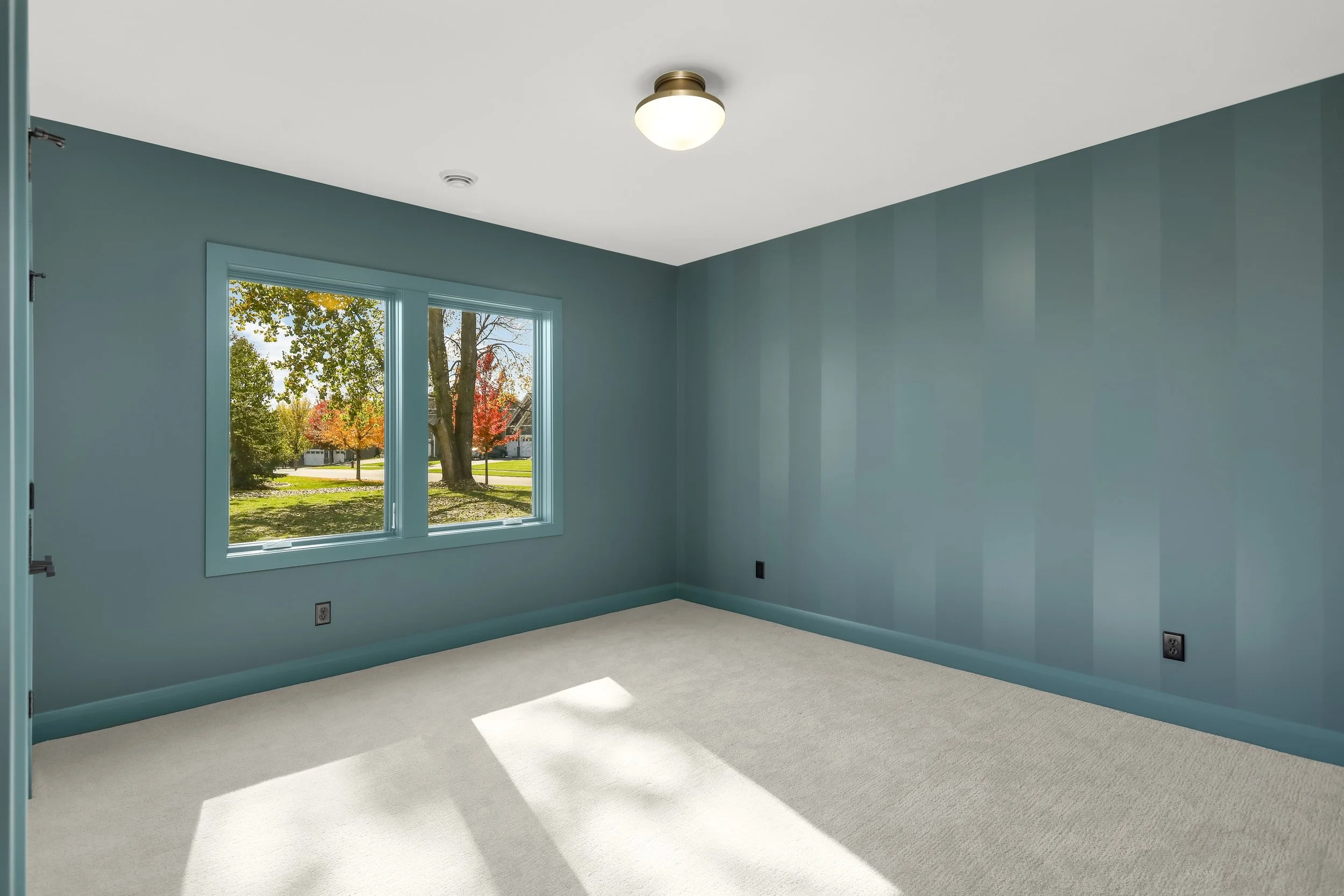 Empty blue-painted room with carpeted floor, double windows, ceiling light, and sunlight streaming in
