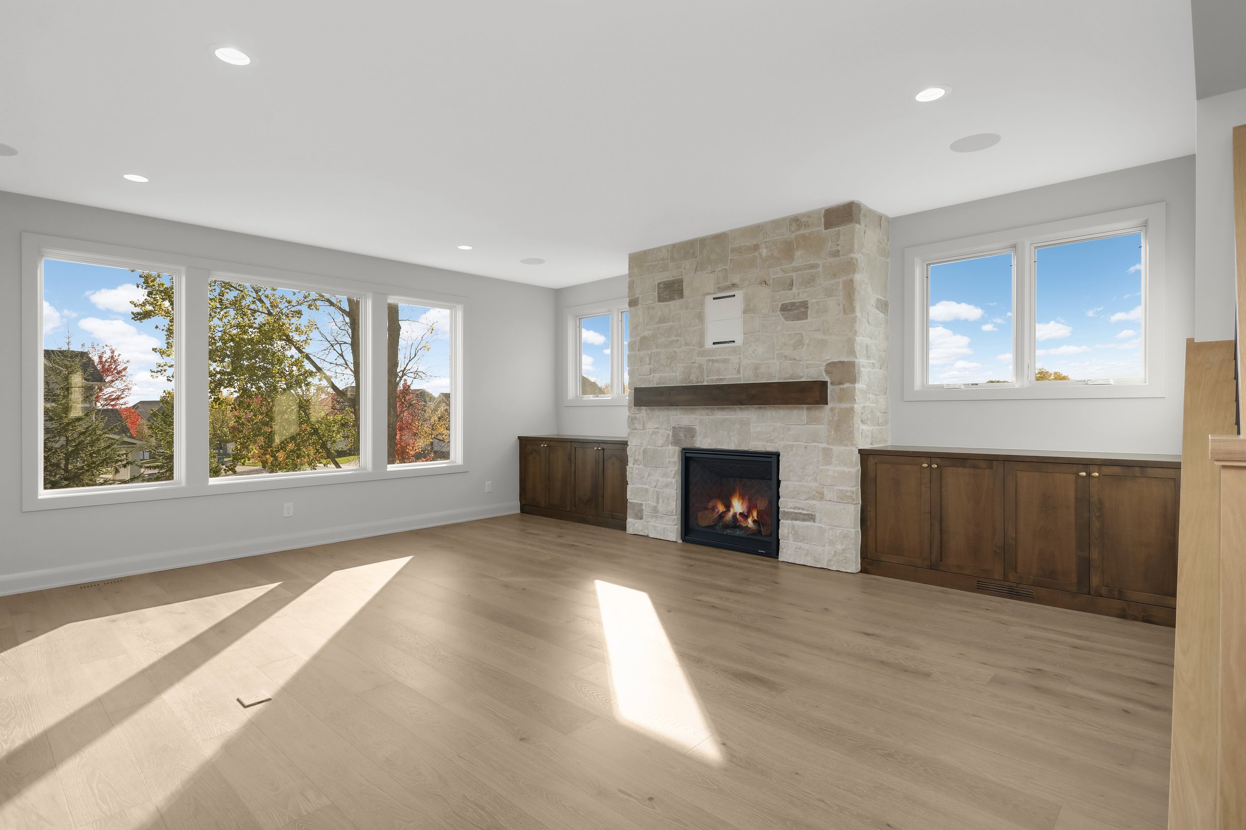 Bright modern living room with a stone fireplace, wood cabinetry, hardwood floors, recessed lighting, and large windows overlooking autumn trees.