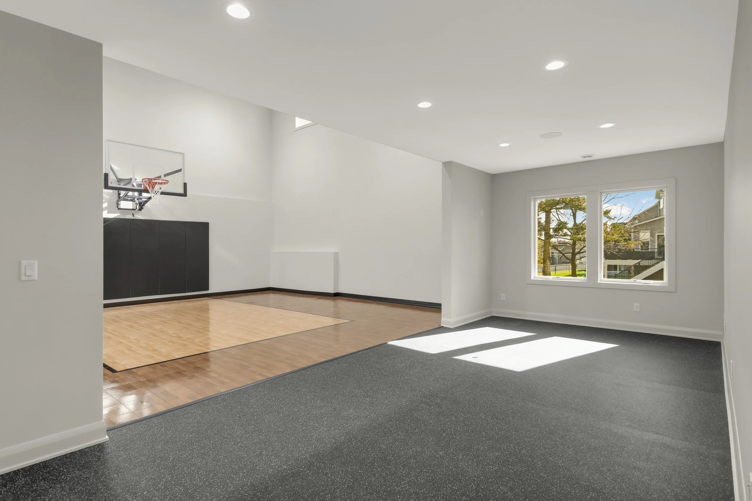 Bright indoor basketball court with hardwood flooring, wall-mounted hoop, black safety padding, windows, and rubber exercise flooring.