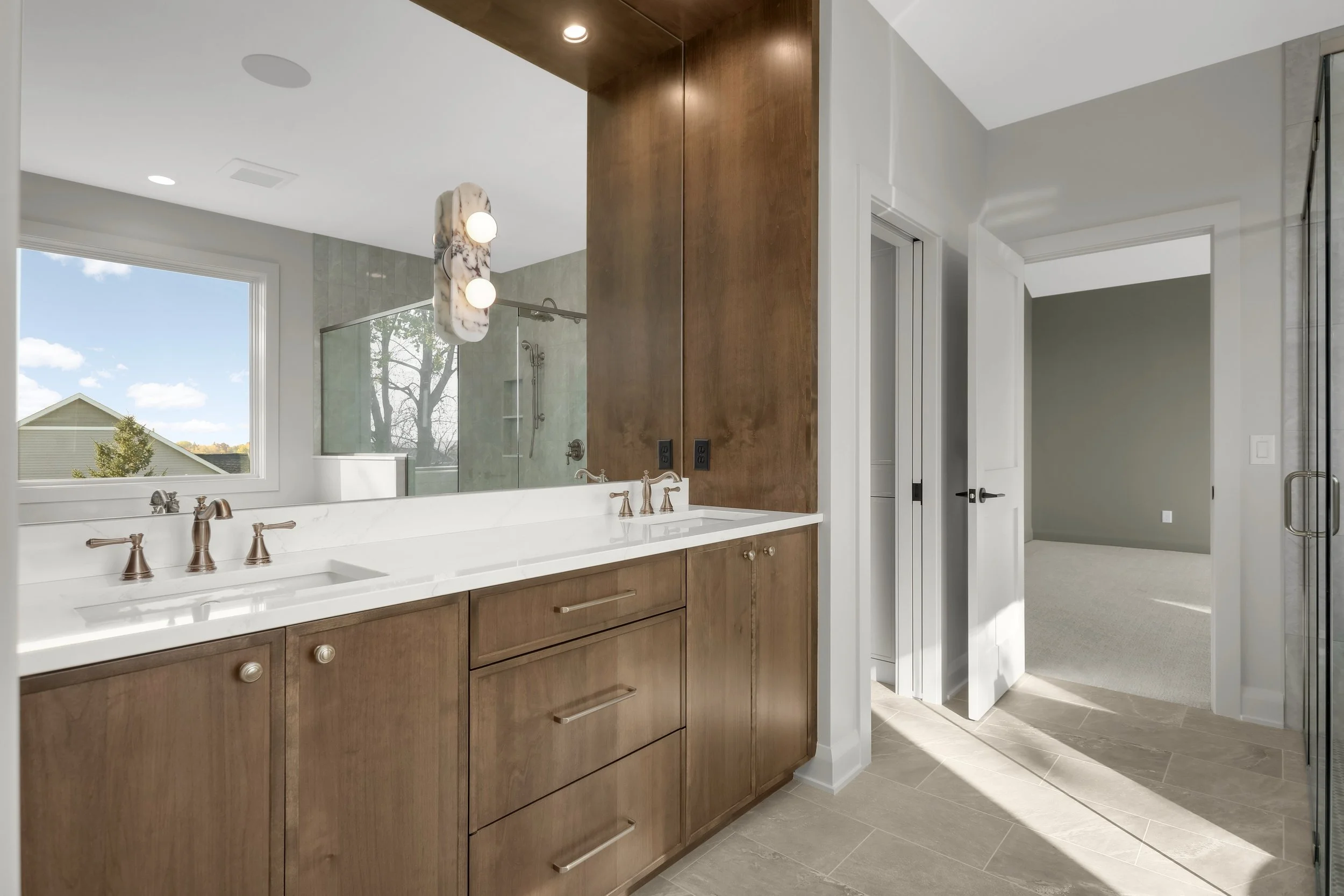 Modern bathroom with double wood vanity, white countertop, large mirror, glass-enclosed shower, and window overlooking a house