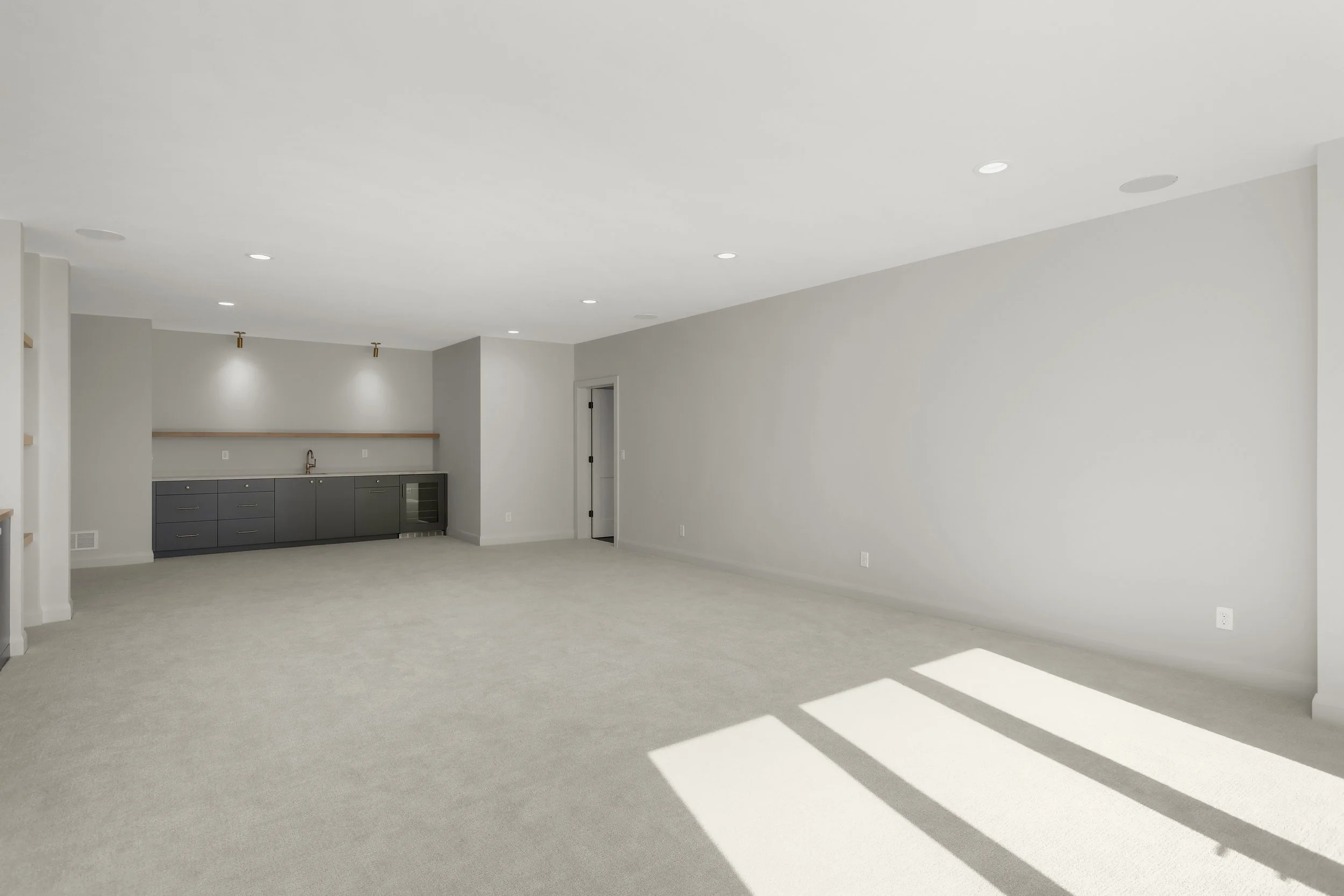 Empty modern living room with carpeted floors, recessed lighting, and a compact kitchenette with dark cabinets and stainless-steel appliances.