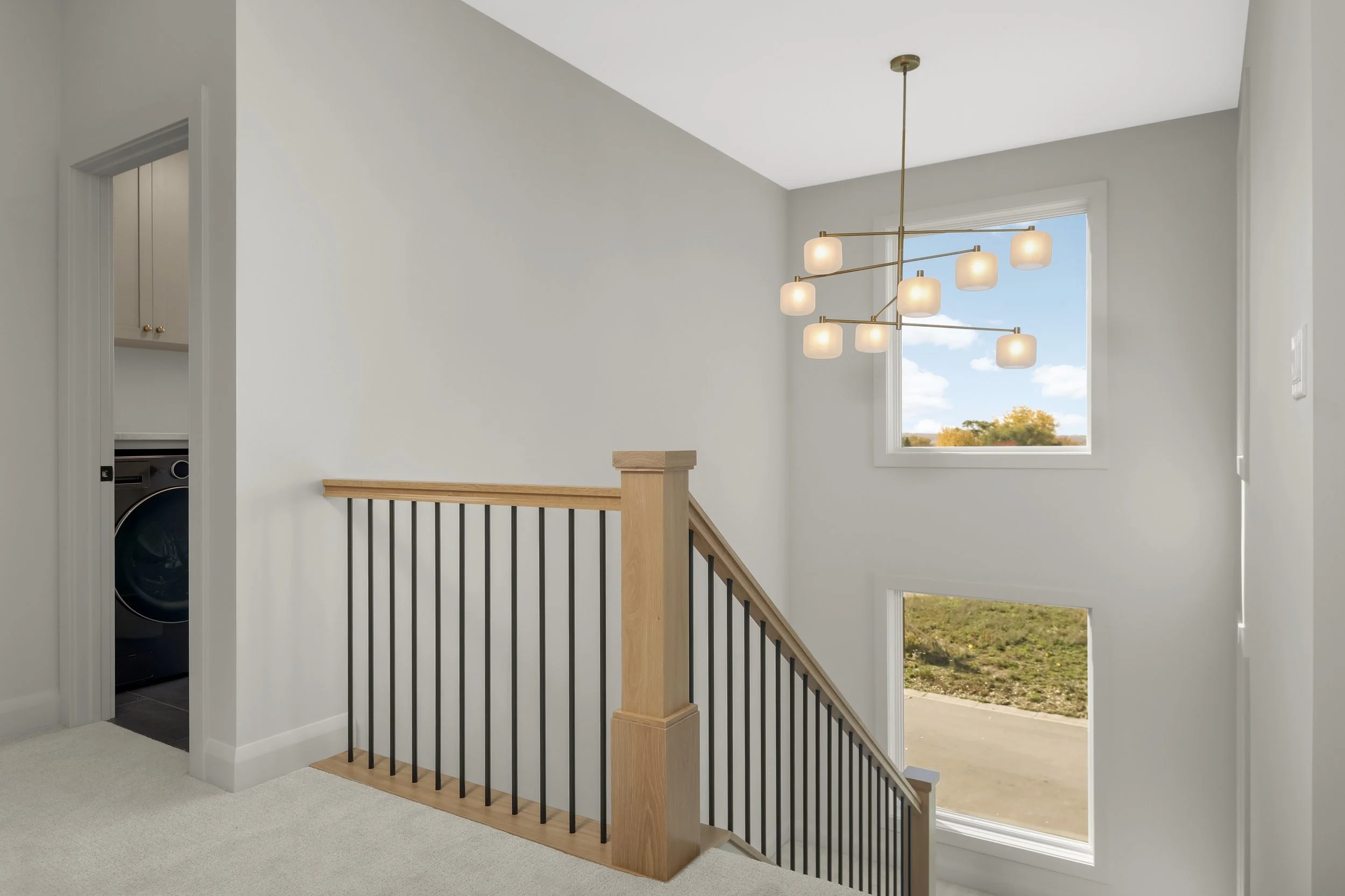 Modern upstairs hallway overlooking a staircase with wood handrails, black metal balusters, large windows, and a contemporary multi-light chandelier
