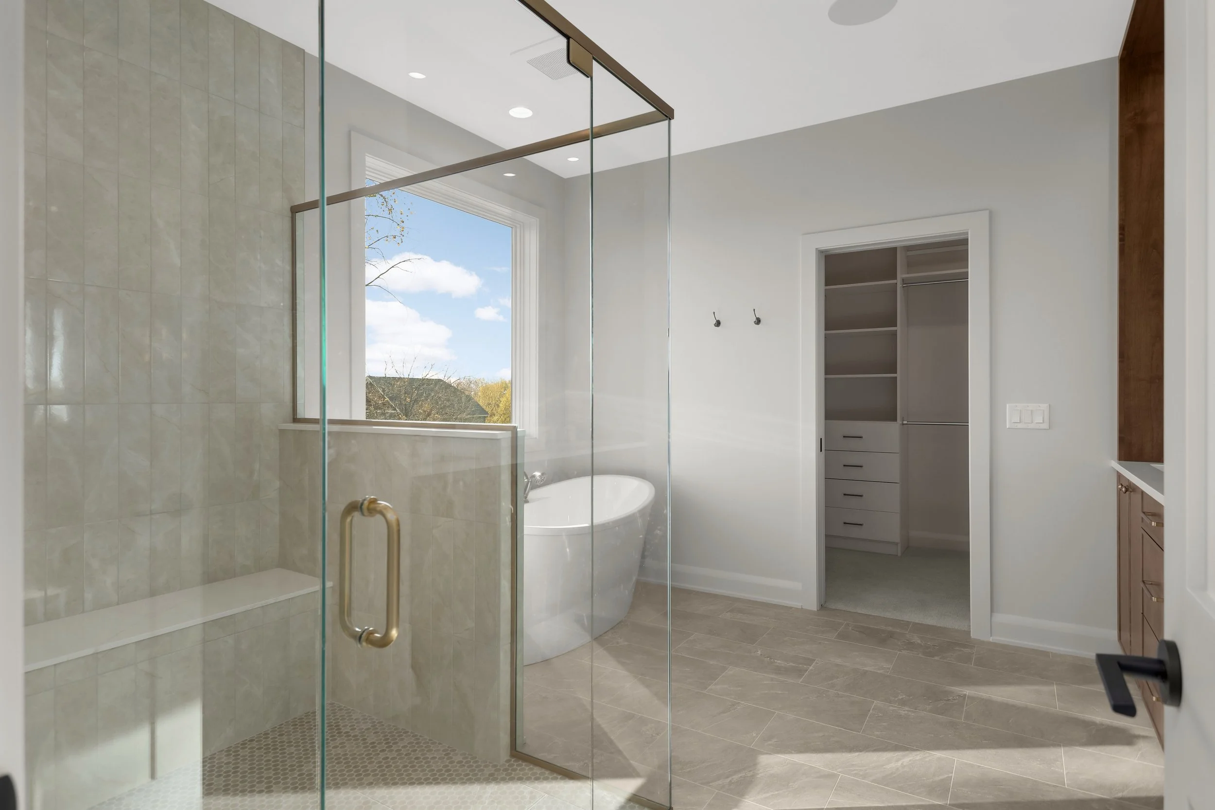 Modern primary bathroom with glass-enclosed tiled shower, freestanding soaking tub, window, walk-in closet, and vanity