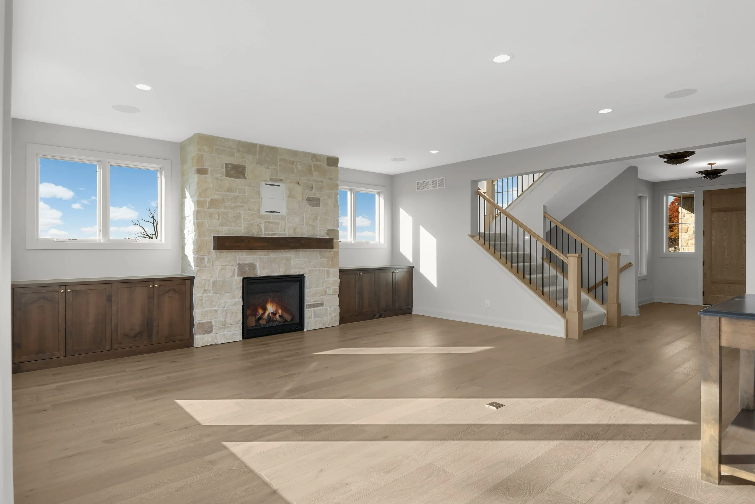 Bright open-concept living room with a stone fireplace, built-in dark wood cabinets, large windows, hardwood floors, and a staircase with black iron railings.