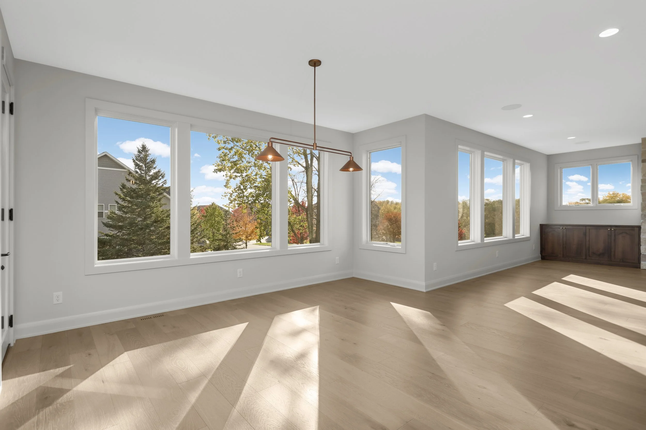 Bright open-concept living room with light wood floors, large windows, pendant lights, and a dark wood sideboard